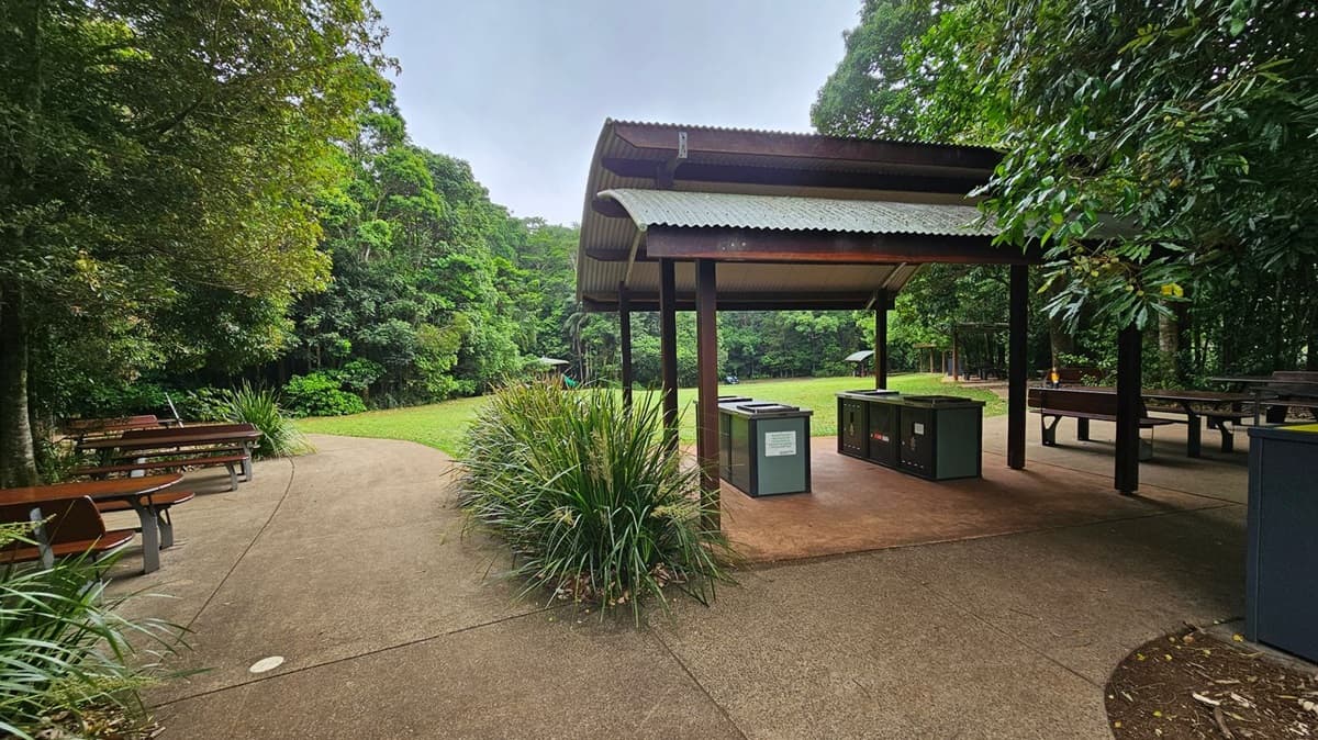 A picnic area with wooden tables and benches on a paved surface. On the left, there's a covered shelter with a barbecue grill. The background features a large grassy field bordered by dense, lush green trees. A small playground with a slide is visible in the distance. Overhead, tree branches with green leaves partially frame the scene.