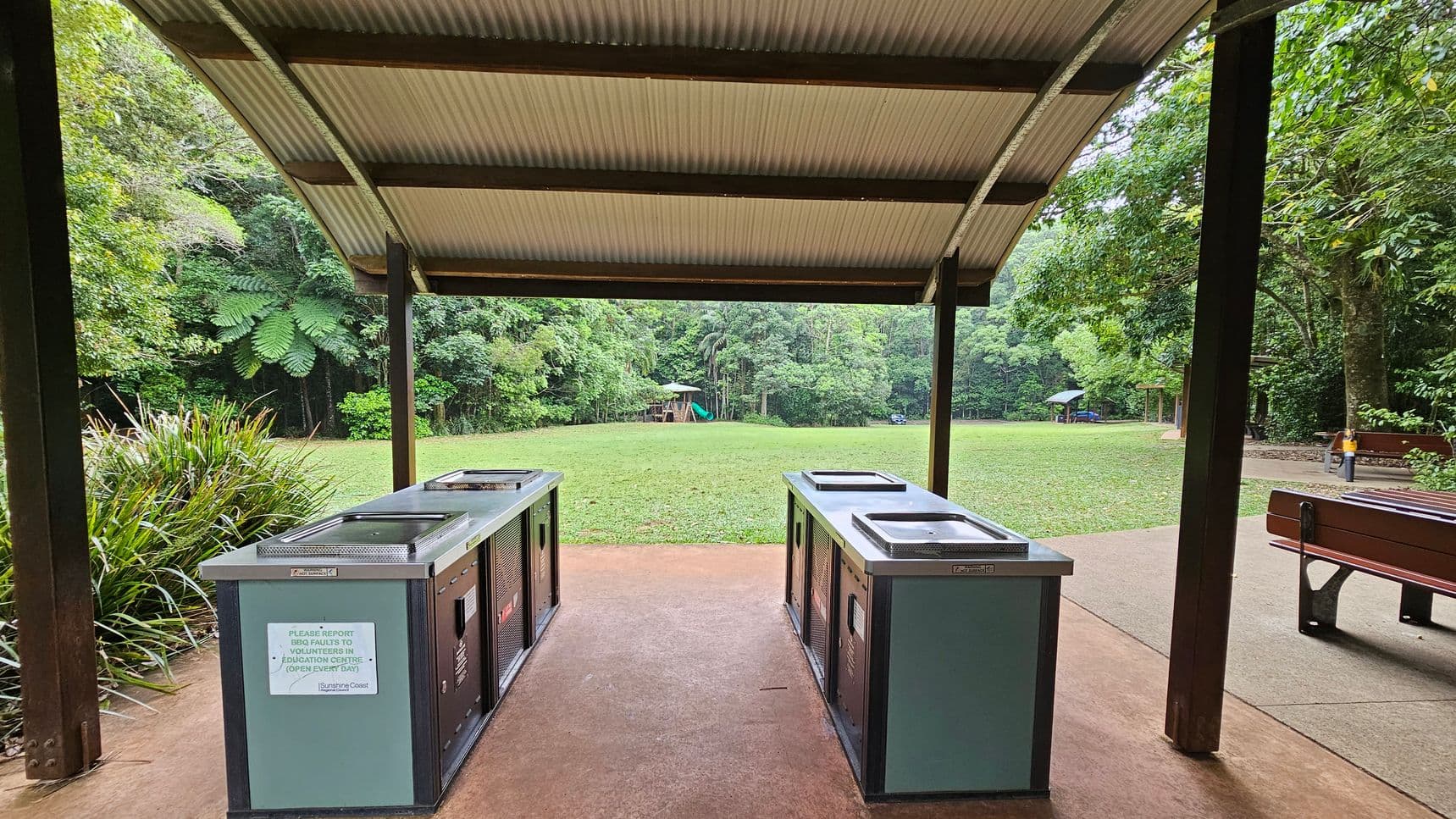 Under a corrugated metal roof, two barbecue stations are positioned on a concrete floor. To the left, dense green foliage and a patch of grass are visible. In the background, a grassy field extends to a playground with a slide. On the right, a wooden bench is partially visible, with more trees and a path leading into the distance.