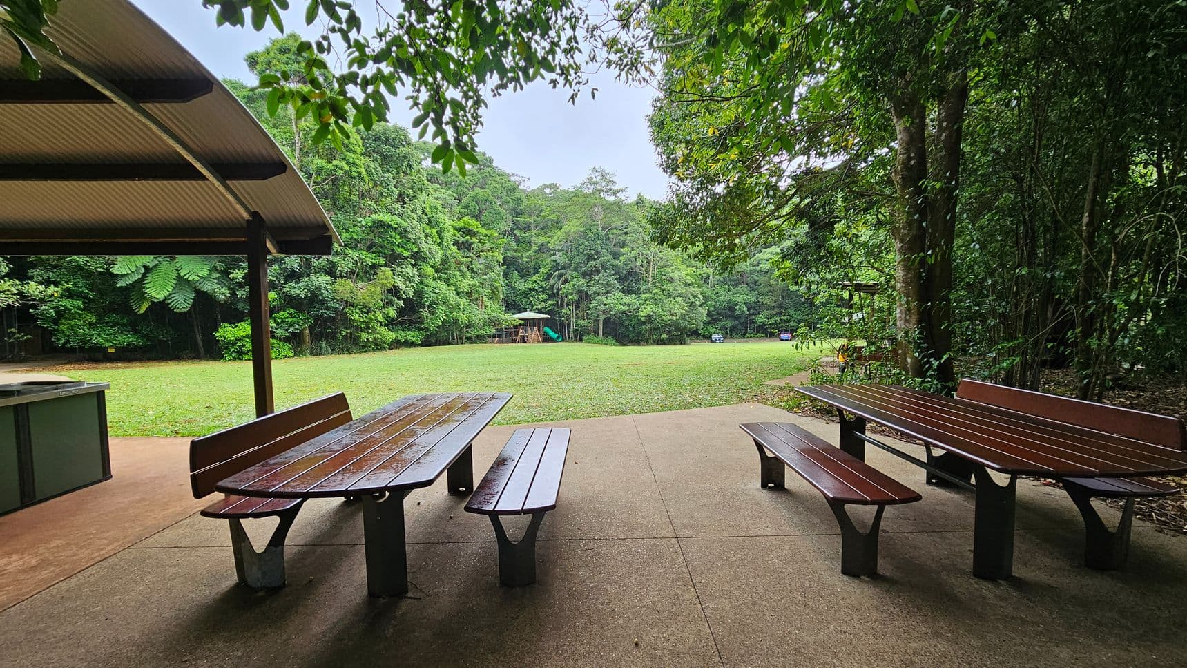 A covered picnic area with wooden tables and benches on a concrete floor. To the left, a metal barbecue grill is visible under a curved roof. In the background, a grassy field extends towards dense green trees. A playground with a slide is visible in the distance, along with a parked car. The scene is surrounded by lush foliage, creating a serene, natural setting.