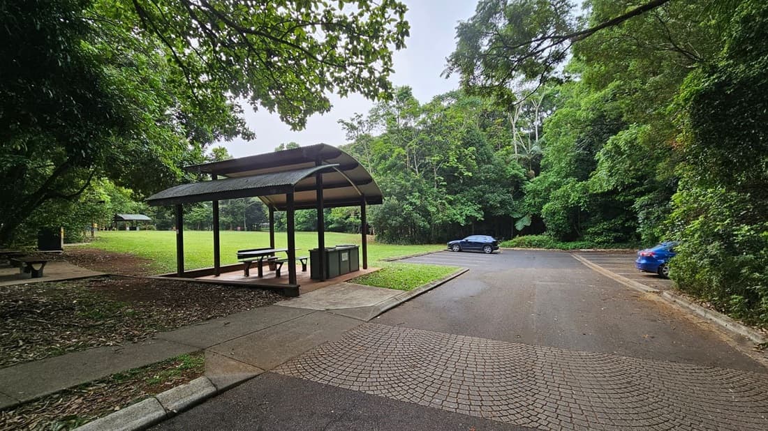 A park scene with a sheltered picnic area on the left, featuring a table and benches under a curved roof. The ground is covered with leaves and grass. In the background, a grassy field is visible, bordered by dense green trees. On the right, there is a paved parking area with two cars, one black and one blue. The foreground shows a textured pavement leading to the parking area.