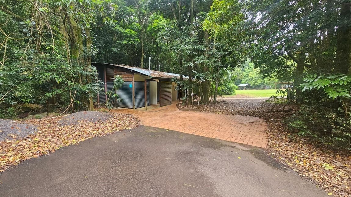 A small corrugated metal building is nestled among dense trees. The ground is covered with fallen leaves, and a paved brick path leads to the building. To the right, there is an open grassy area with more trees in the background. The foreground features a dark asphalt surface.
