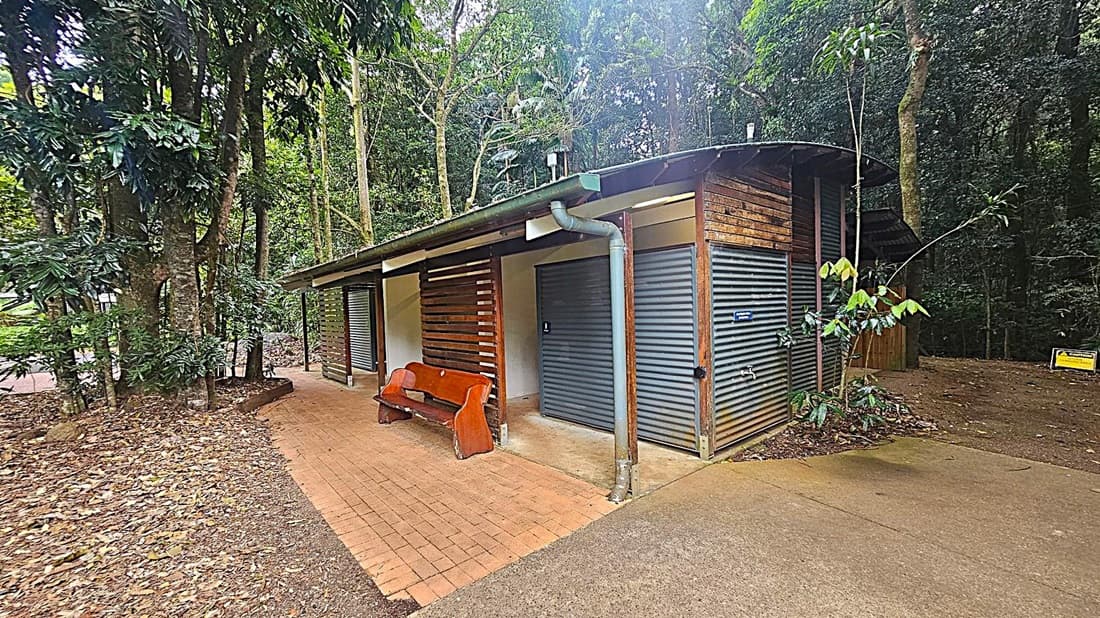 A small public restroom building with corrugated metal doors and wooden slats. A wooden bench is positioned in front of the building on a brick-paved area. The structure is surrounded by dense forest with tall trees and lush green foliage. A concrete path leads to the building, and a caution sign is visible on the right side near the trees. The roof is slightly curved with a gutter system. The ground is covered with fallen leaves.