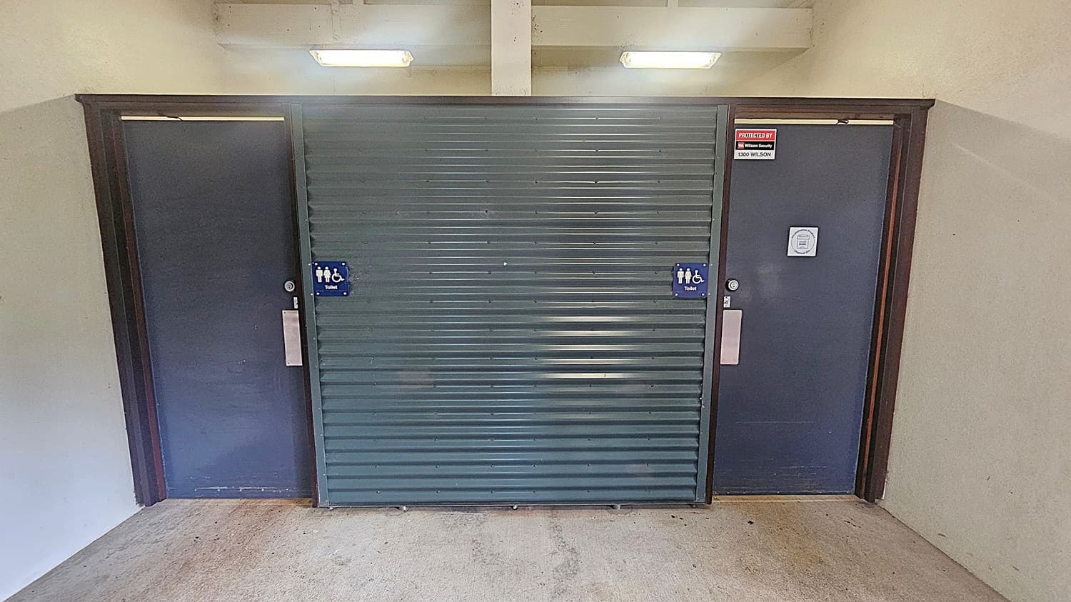 A grey metal roller door is in the centre, flanked by two dark blue doors on either side. Both doors have signs indicating accessible toilets. The right door has a security notice above it. The floor is concrete, and the ceiling has two fluorescent lights. The walls are light-coloured.