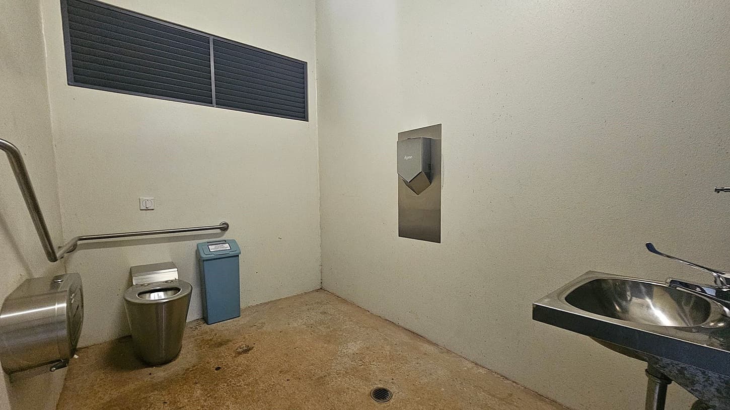A public toilet with a stainless steel toilet on the left, accompanied by a toilet paper dispenser and a grab rail. Next to it is a blue sanitary bin. On the right, a stainless steel sink with a tap is visible. A hand dryer is mounted on the wall above the sink. The floor is concrete with visible stains, and the walls are light-coloured. A black vent is located high on the left wall.