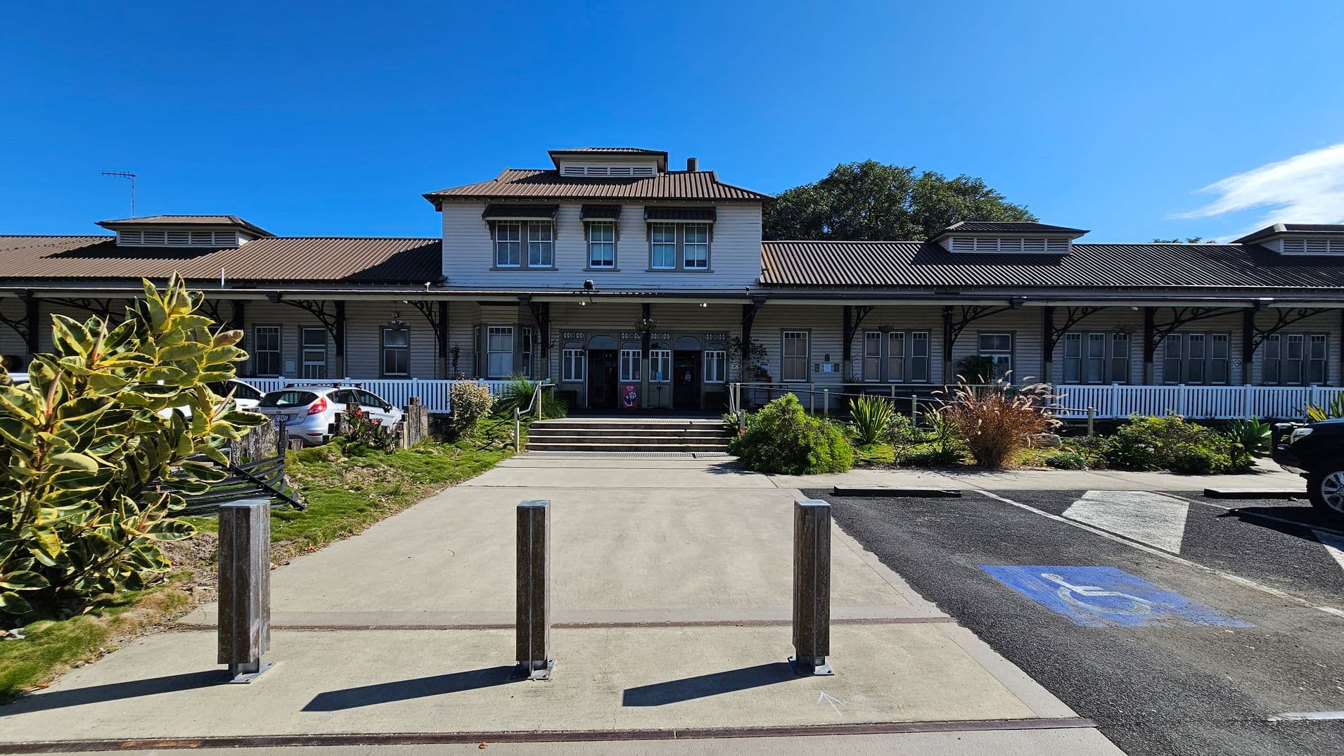 A large, two-storey building with a brown, corrugated roof and multiple windows. The entrance features a set of steps leading up to double doors. In front, there are three metal bollards on a paved path. To the left, a small garden with shrubs and a parked white car. On the right, a marked disabled parking space on the tarmac. The building is surrounded by greenery, including bushes and trees.