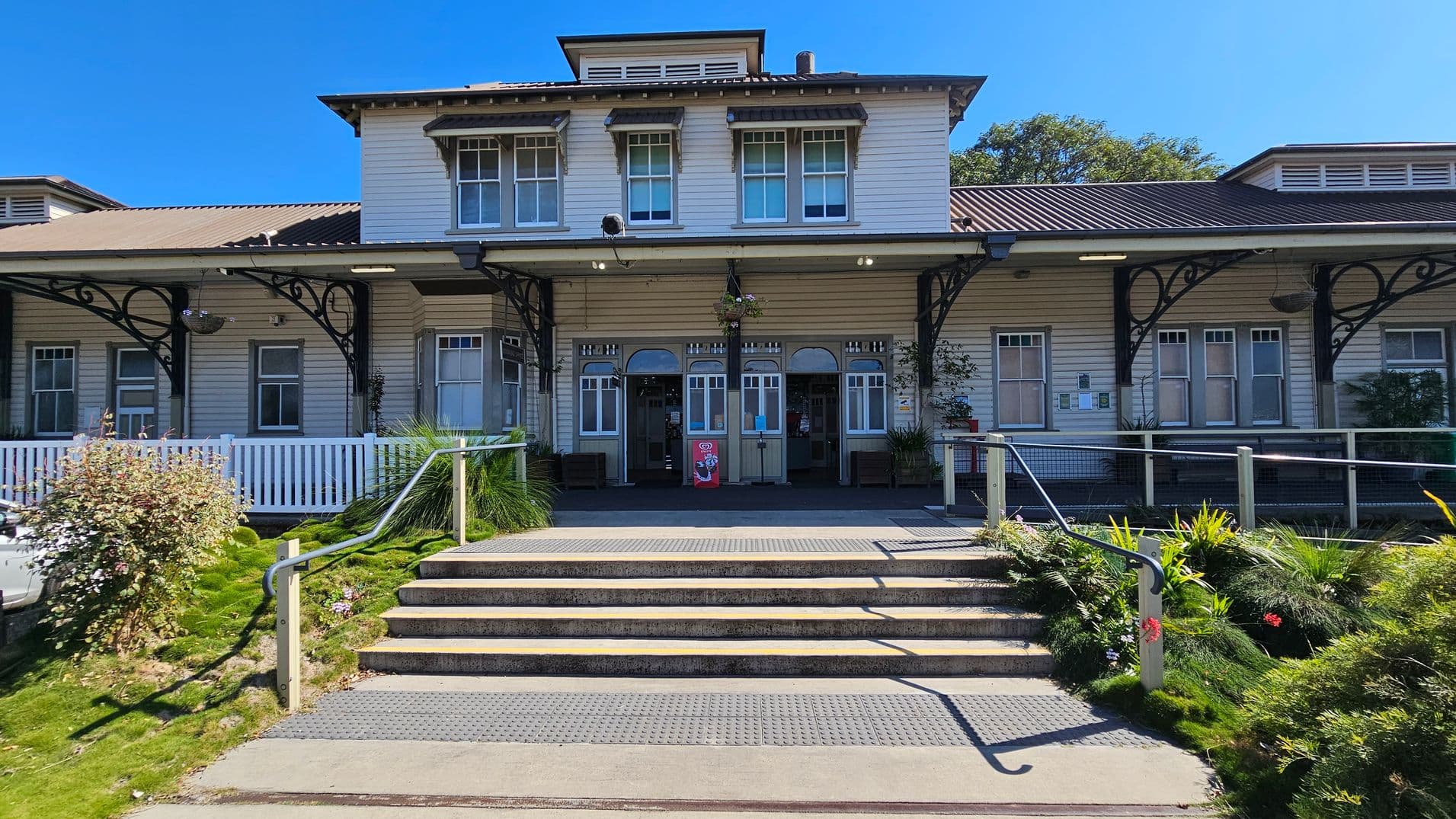 A historic building with a symmetrical facade, featuring a central entrance with double doors and large windows on either side. The structure is adorned with decorative ironwork and a pitched roof. A set of steps leads up to the entrance, flanked by greenery and a white picket fence on the left. The building is surrounded by well-maintained gardens, and a ramp with a handrail is visible on the right. The exterior is painted in a light colour with dark trim.