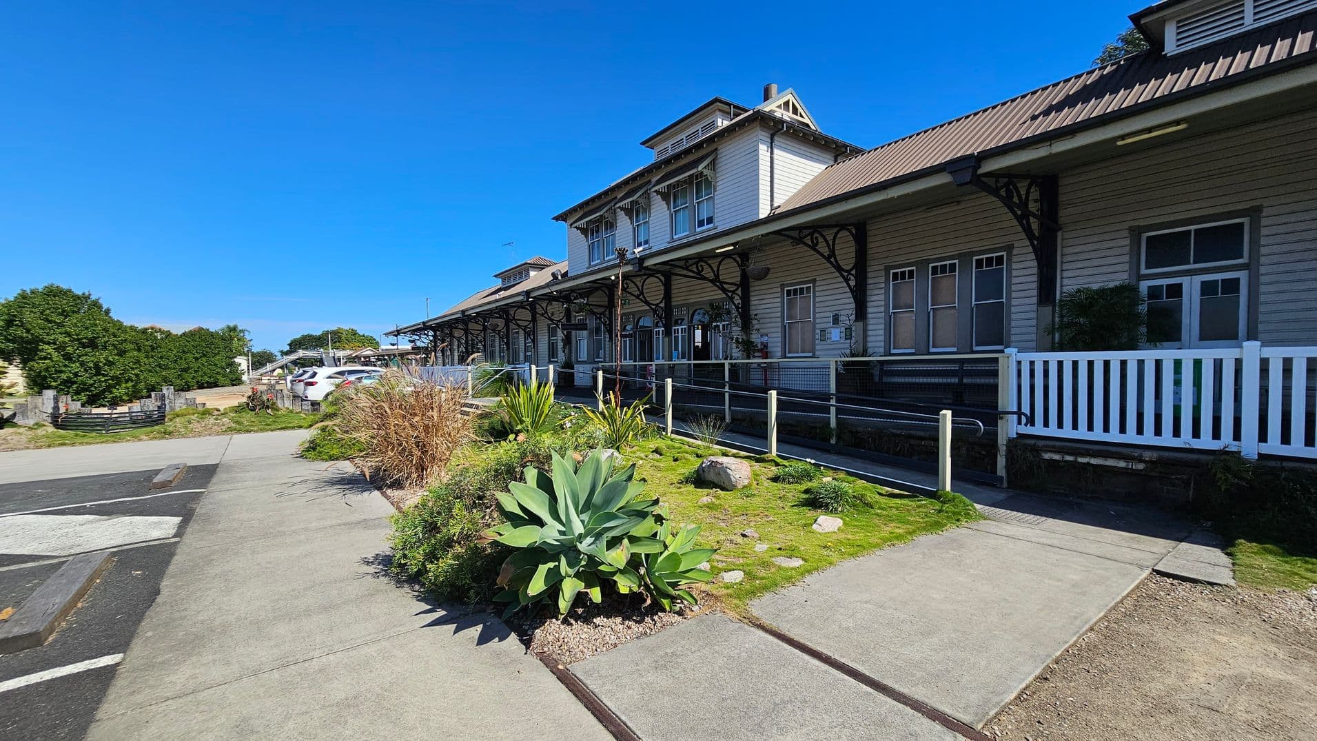 A historic wooden building with a corrugated metal roof and decorative ironwork supports a covered walkway. The structure has multiple windows and a white picket fence. In the foreground, there is a garden with various plants and a stone path. To the left, a paved area leads to a car park with several vehicles. Bushes and trees are visible in the background. The sky is clear and blue.