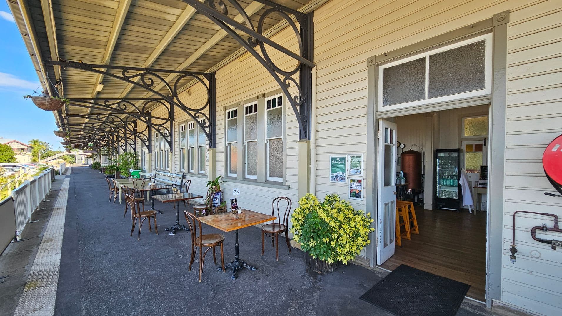 A covered outdoor area with a row of tables and chairs along a wooden building. The building has large windows and decorative metal brackets supporting the roof. To the right, an open door leads inside, revealing a fridge and stools. A potted plant sits near the entrance. The ground is paved, and a railing runs along the left side, with a view of parked cars and trees in the background. Hanging baskets are attached to the roof supports.