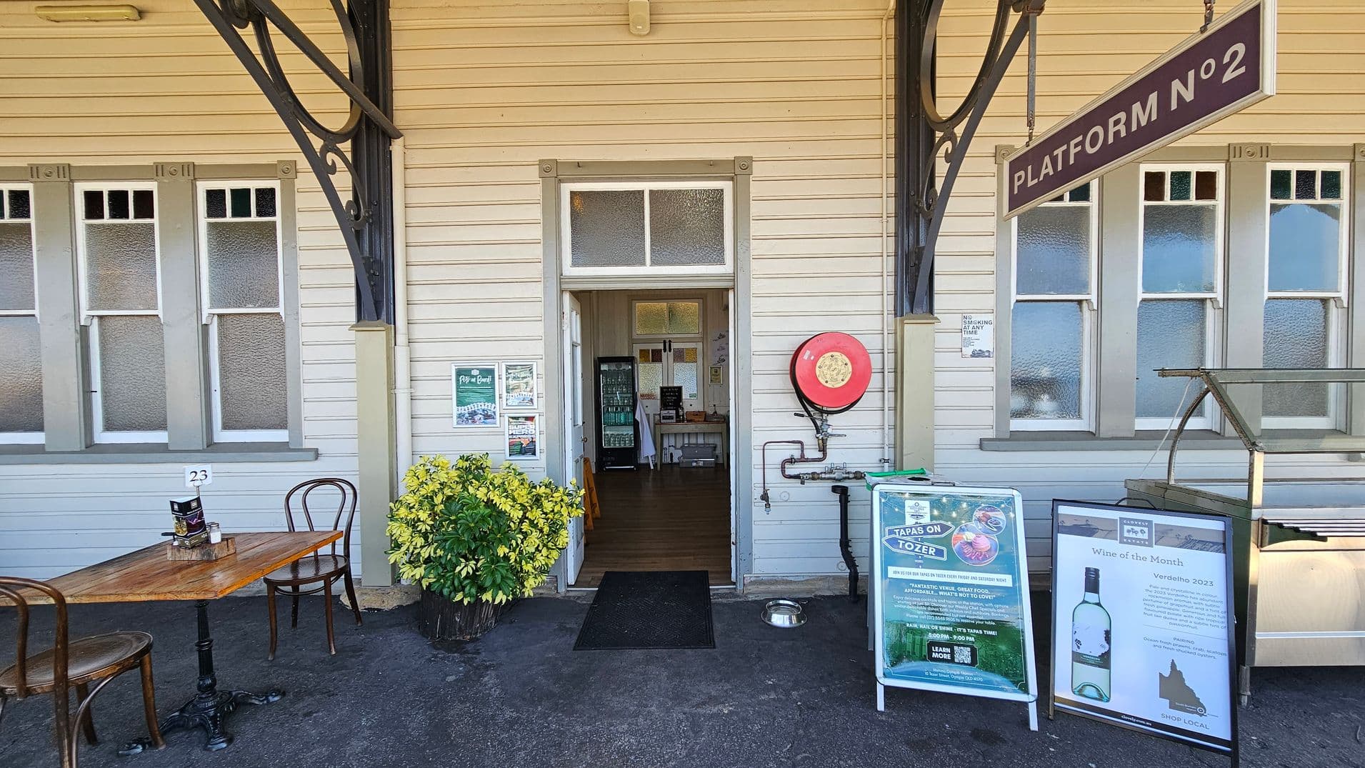 A wooden building with cream-coloured walls and frosted windows. In the centre, a door leads inside, flanked by a red fire hose reel. To the left, a small table with two chairs and a potted plant. Above, a sign reads "PLATFORM N°2". On the right, two signs advertise "Tapas on Tozer" and "Wine of the Month". The floor is dark and textured, and the ceiling is not visible.