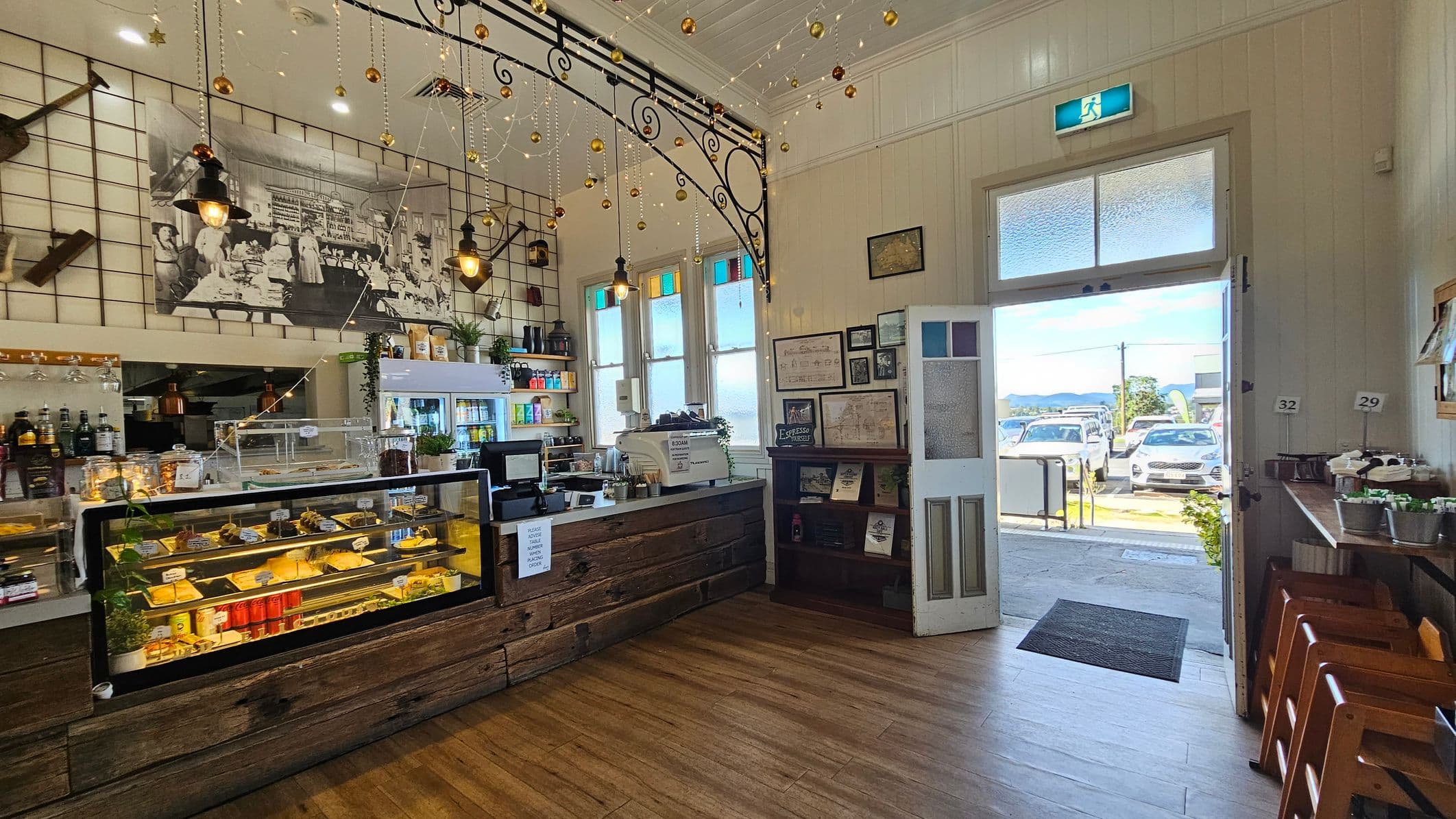 Inside a café with wooden flooring and a high ceiling. On the left, a counter with a glass display case filled with pastries and drinks. Above, a large black-and-white photo and hanging lights with gold decorations. In the middle, a counter with a cash register and shelves stocked with various items. On the right, an open door leading outside, with sunlight streaming in. Near the door, a small table with condiments and stools. The walls are adorned with framed pictures and a stained glass window.