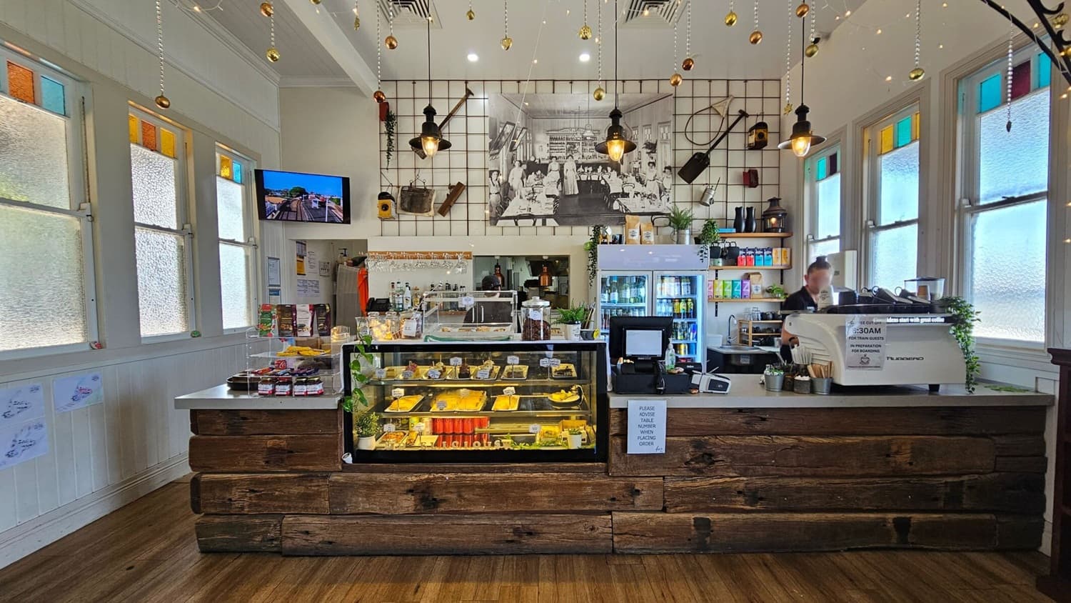 A café interior with a rustic wooden counter in the foreground, displaying pastries and snacks in a glass case. To the right, a barista operates a coffee machine. Behind the counter, a fridge stocked with drinks and shelves with various items. The back wall features a large black-and-white photo and vintage tools. The ceiling has hanging lights and decorations. The left and right walls have windows with coloured panes, and the floor is wooden.