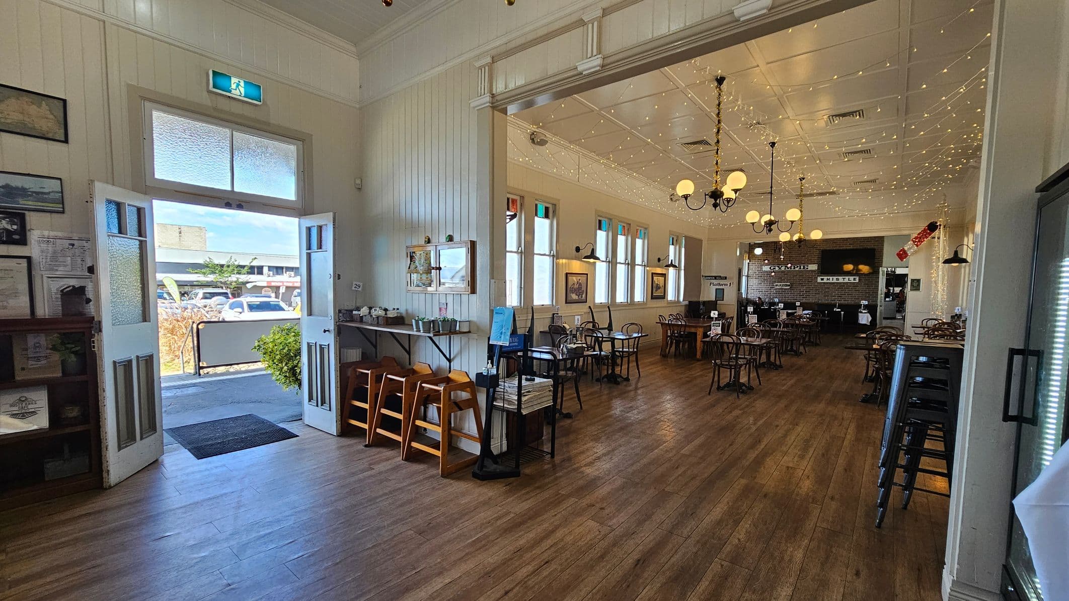 A spacious café interior with wooden flooring and high ceilings adorned with string lights. On the left, a set of double doors with frosted glass panels opens to the outside, where cars are visible. Inside, there are wooden chairs and tables arranged neatly. The right side features a bar area with stools and a sign reading "Platform 1" and "Whistle". The walls are decorated with framed pictures, and there are multiple windows allowing natural light to enter.
