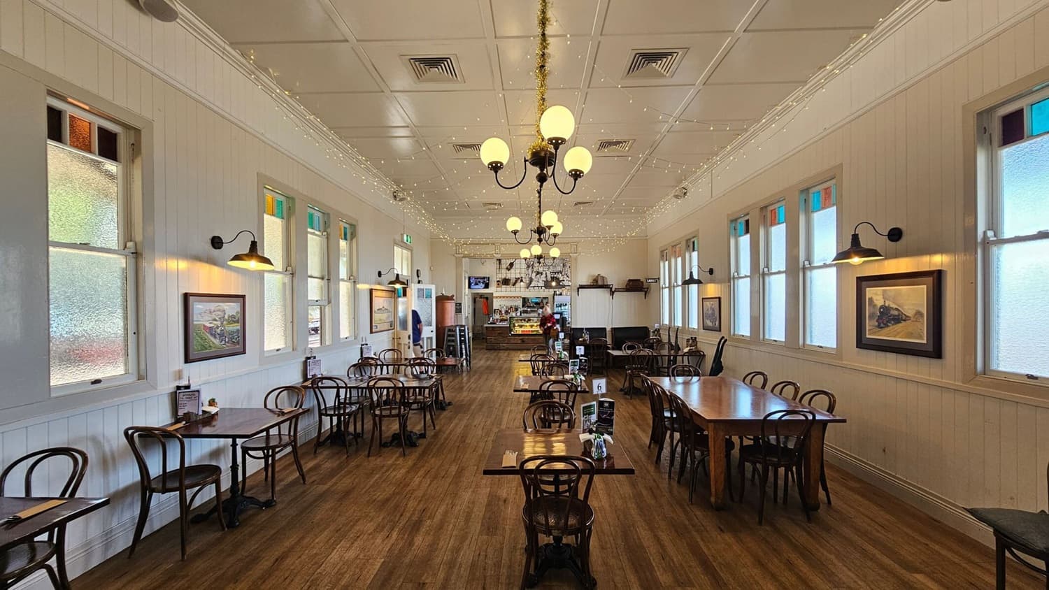 A spacious café interior with wooden flooring and a high, white panelled ceiling adorned with chandeliers and string lights. On both sides, there are large windows with frosted glass and colourful panes at the top. The walls are decorated with framed pictures. Wooden tables and chairs are neatly arranged throughout the room. At the far end, a counter with a display case and a person behind it is visible, along with a small television mounted on the wall.