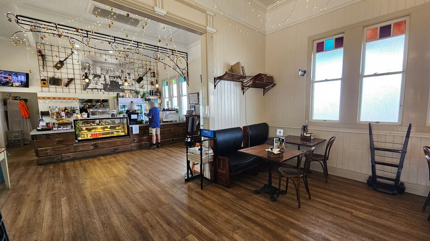 A cosy café interior with wooden flooring. On the left, a counter with a glass display case filled with pastries and a barista serving a customer. Above the counter, decorative lights and musical instruments hang on a grid. In the middle, a black and white photo is displayed. On the right, a seating area with a table, chairs, and a bench. Two large windows with coloured panes are on the wall, and a stack of newspapers is on a rack near the table.