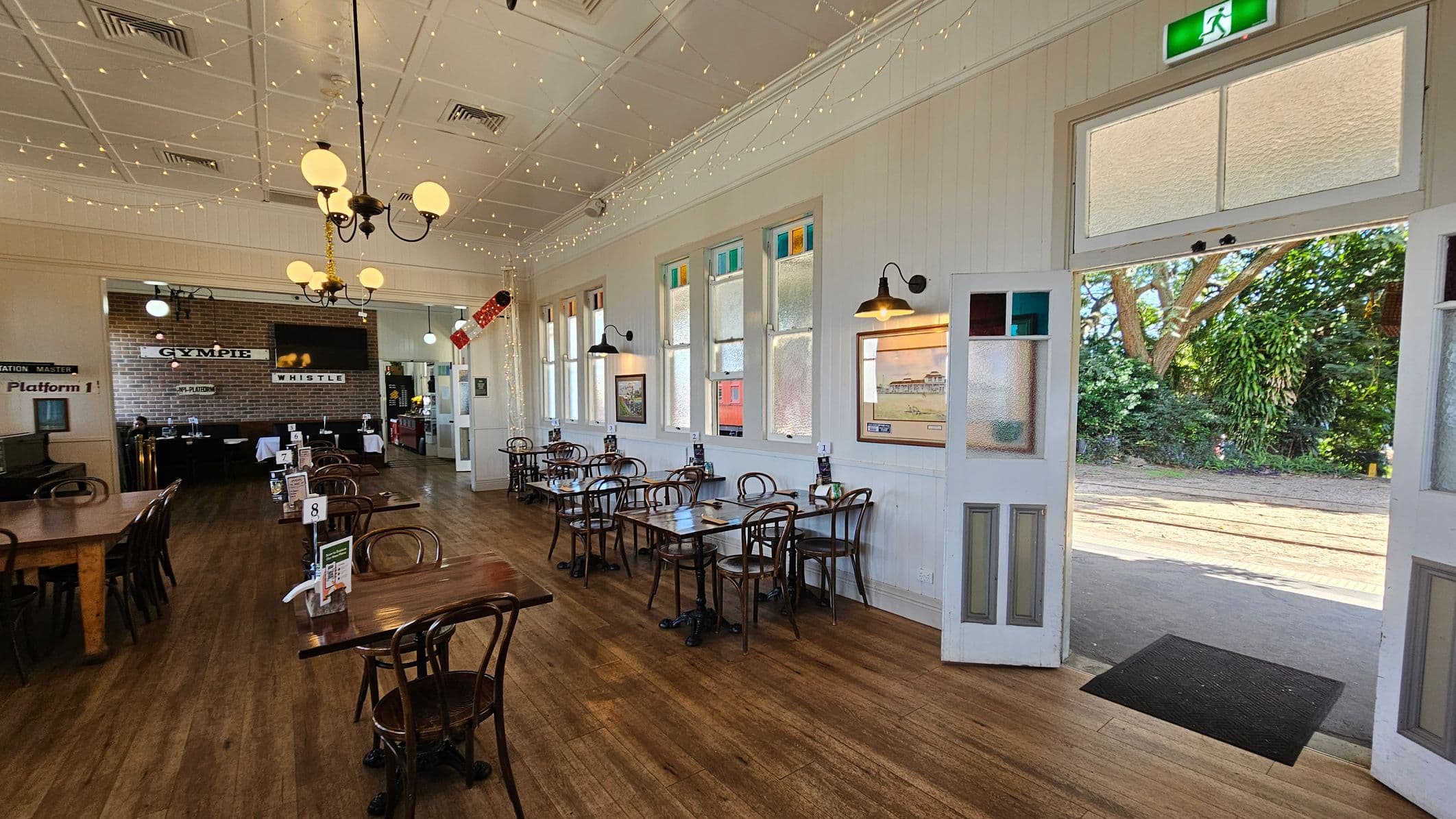 A spacious café interior with wooden flooring and a white panelled ceiling adorned with string lights. On the left, there are wooden tables and chairs, with a brick wall featuring signs and a TV. The right side has a row of windows and more tables with chairs. An open door leads outside, revealing greenery and a tree. A chandelier with globe lights hangs from the ceiling, and wall-mounted lamps provide additional lighting. An exit sign is above the door.