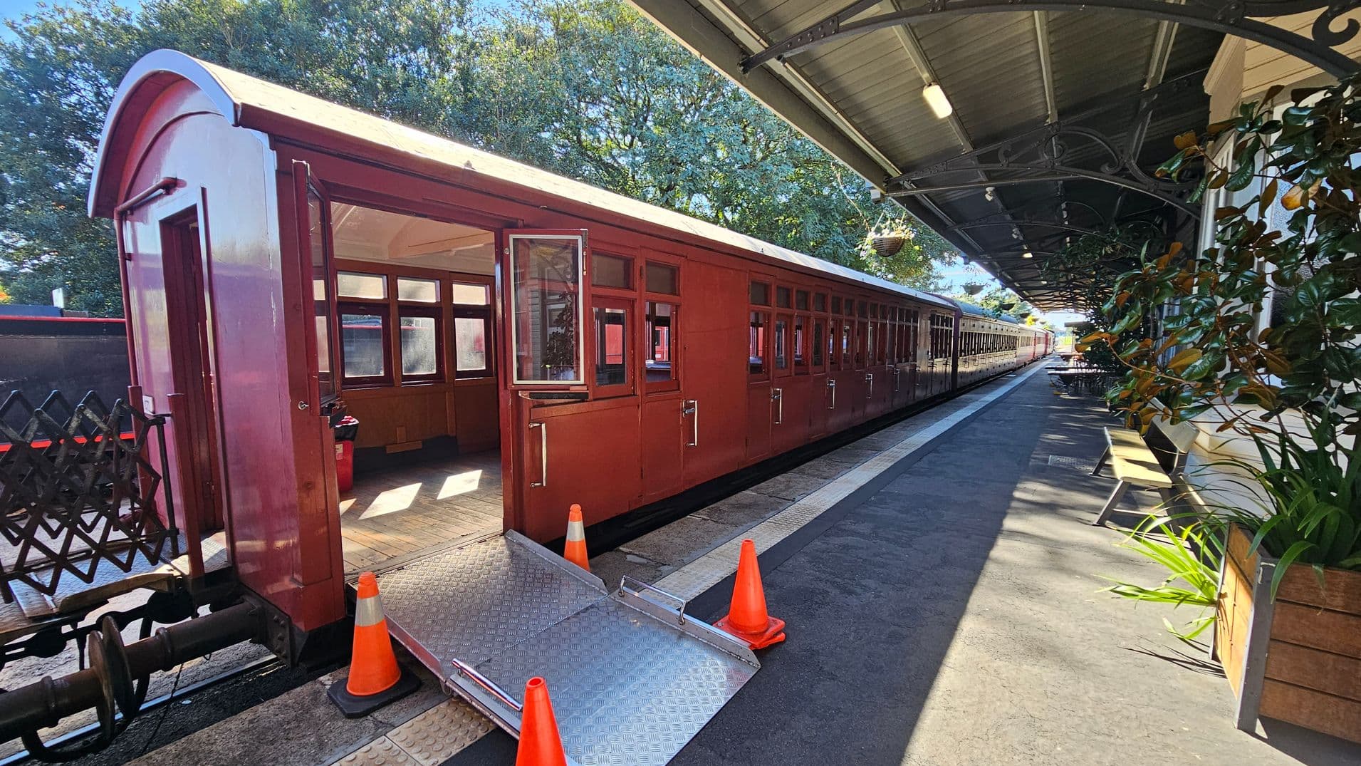 A vintage red train is stationed on the left, with an open door and a metal ramp leading to the platform, marked by orange cones. The platform on the right features a covered area with a green roof, decorative metal supports, and hanging baskets. Wooden benches and potted plants line the platform, which extends into the distance. Trees are visible in the background, providing a natural setting.