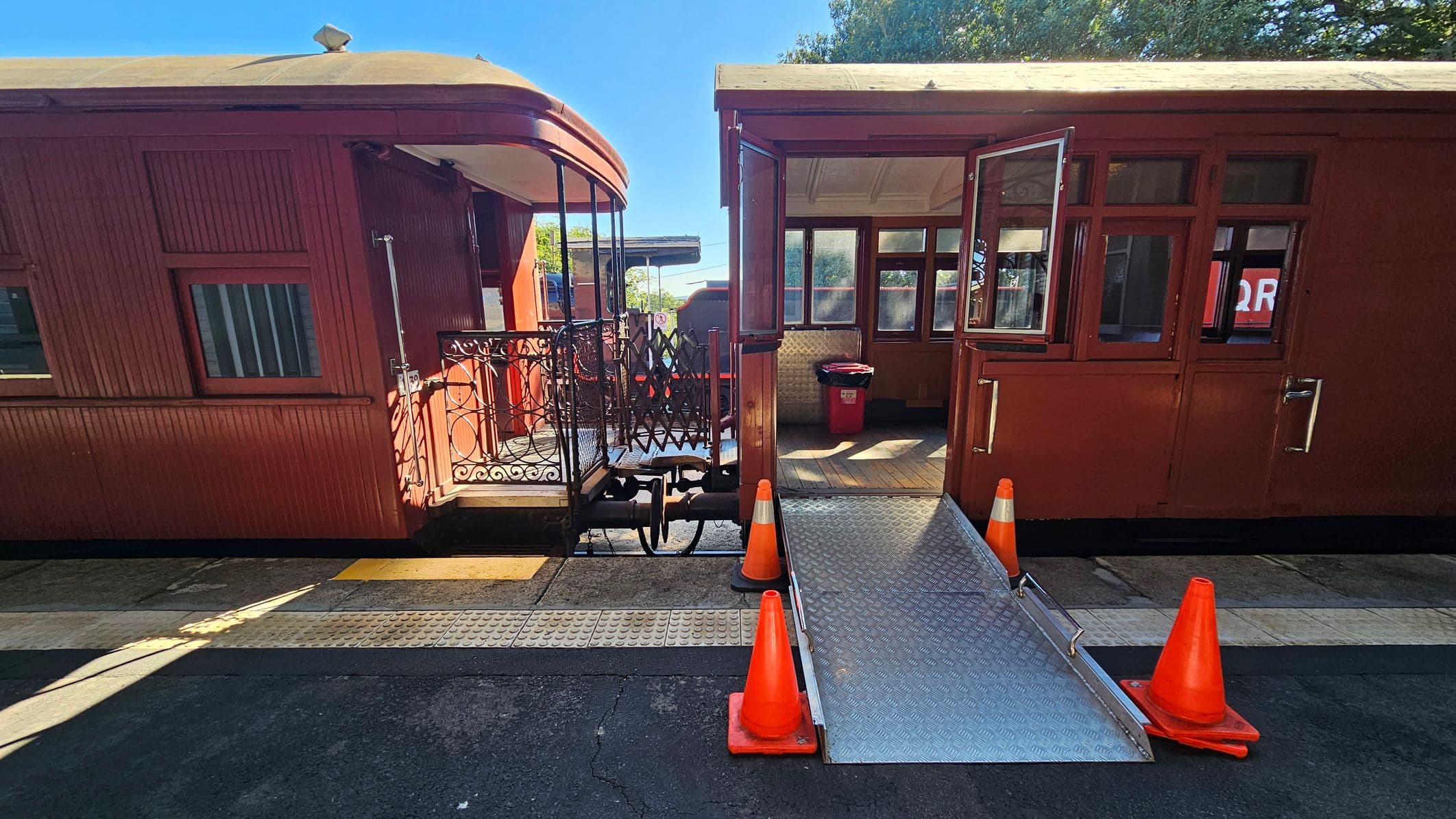 Two vintage train carriages are connected by a small platform with ornate metal railings. On the right, a metal ramp leads into the open door of one carriage, flanked by orange traffic cones. The carriage interiors are visible through the windows, with a red bin inside. The ground is a mix of textured tiles and asphalt. Trees and a structure are visible in the background.