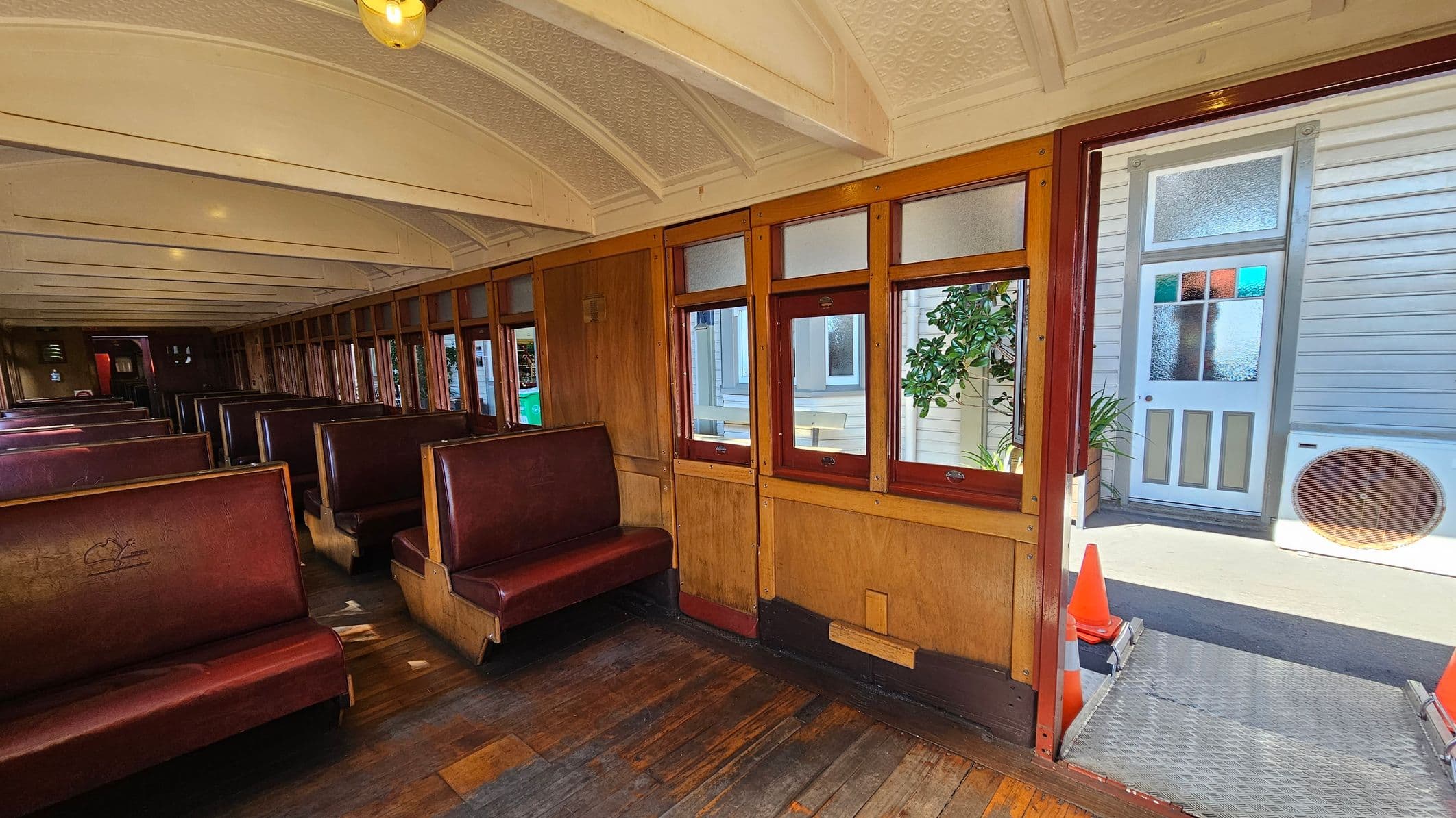 Inside a vintage train carriage with wooden floors and a curved, patterned ceiling. On the left, rows of maroon leather bench seats line the carriage. The right side features wooden panelled walls with windows, through which a building with a white door and frosted glass is visible. An orange traffic cone is near the open doorway, leading to a metal ramp outside. A potted plant and an air conditioning unit are also visible outside.