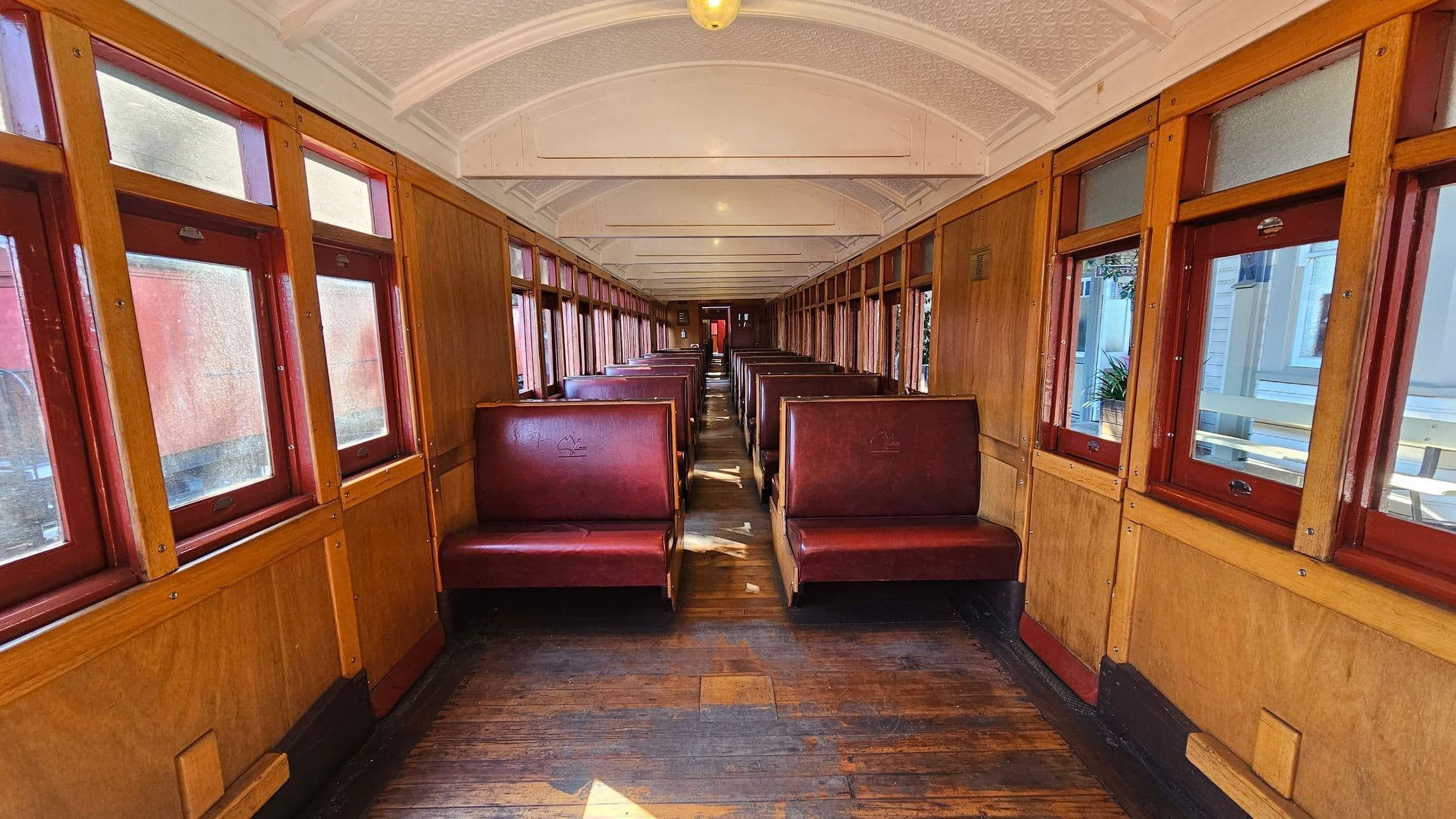 Inside a vintage train carriage with wooden panelling and red leather seats arranged in pairs along both sides. Large windows with wooden frames line the walls, allowing natural light to illuminate the interior. The floor is made of worn wooden planks, and the ceiling is arched with a textured white finish. A single light fixture hangs from the ceiling, and a door is visible at the far end of the carriage.