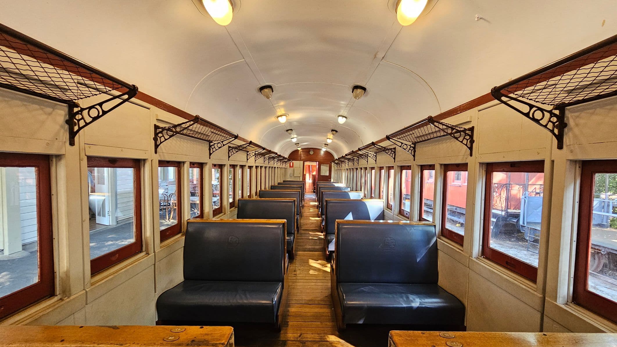 Inside a vintage train carriage with wooden floors and a curved white ceiling. Rows of blue upholstered bench seats are on either side, with wooden tables in front. Above the seats are metal luggage racks. The walls have a series of rectangular windows with wooden frames, allowing light to stream in. Ceiling lights are evenly spaced along the aisle. At the far end, a wooden door is visible.
