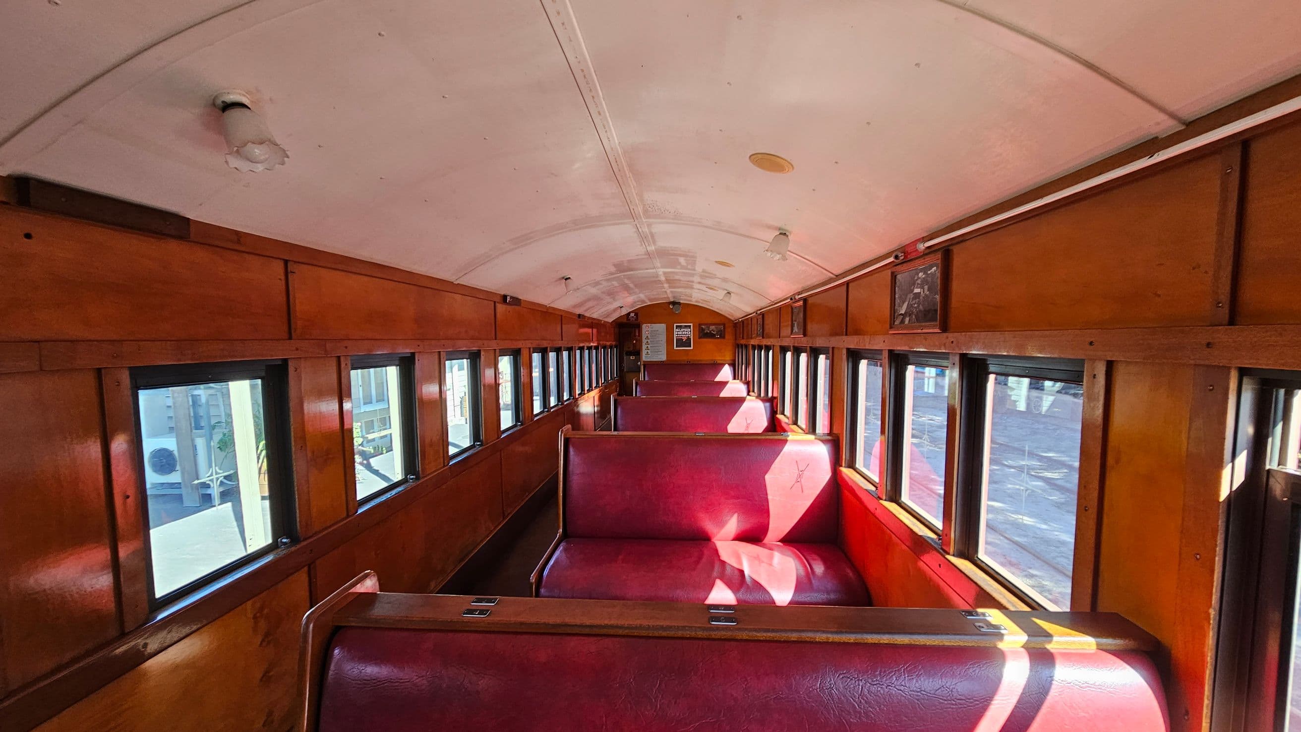 Interior of a vintage train carriage with wooden panelling and red leather bench seats. The left and right sides have a series of rectangular windows allowing natural light to illuminate the space. The ceiling is curved and painted white, with several small light fixtures. At the far end, there are posters and a framed picture on the walls. The floor is dark, contrasting with the warm tones of the wood and seats.