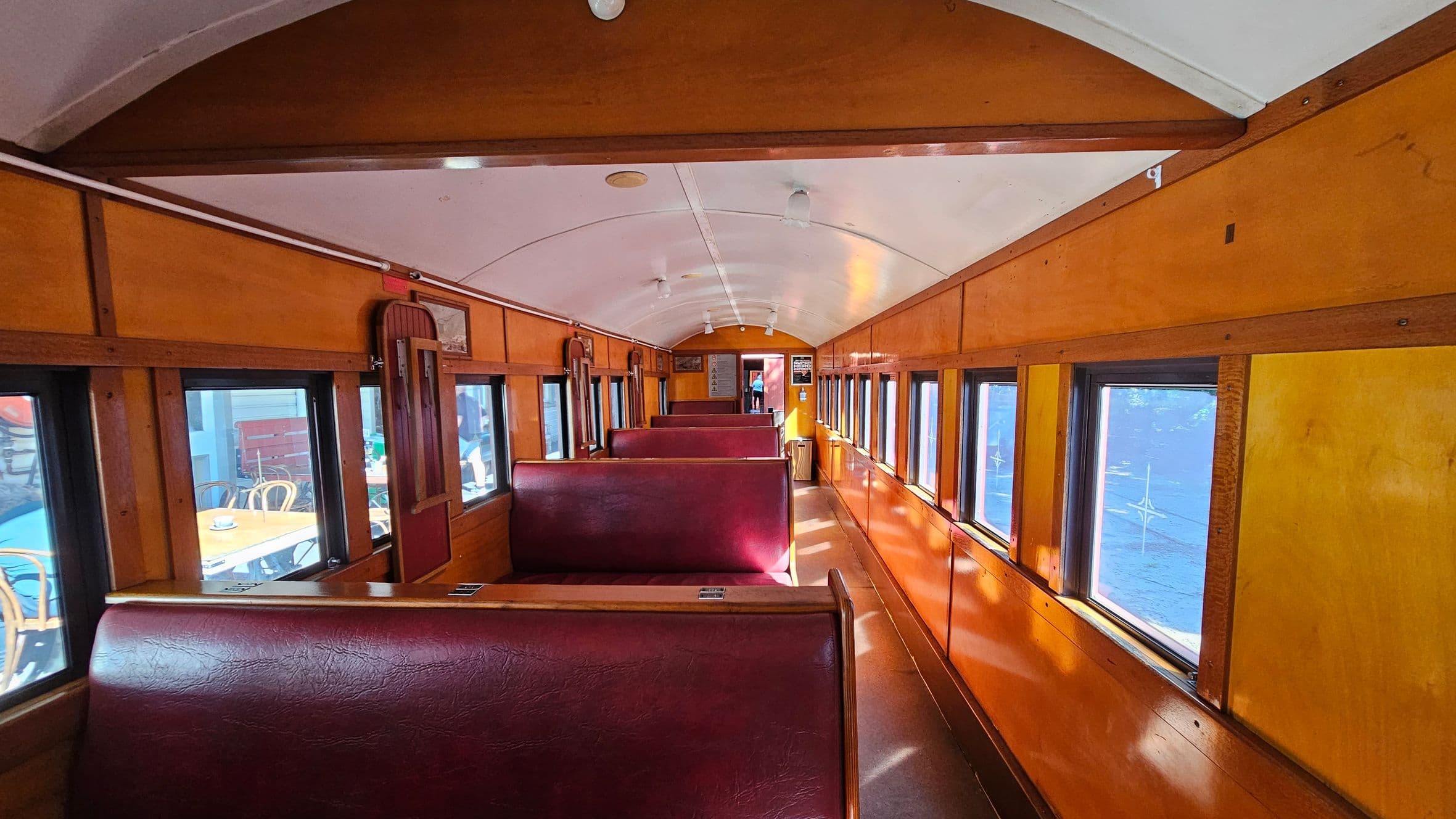 Inside a vintage train carriage with wooden panelling and a curved white ceiling. The floor is a warm brown. On both sides, there are rows of maroon leather bench seats. Large windows line the walls, allowing natural light to illuminate the interior. A small wooden door is visible towards the back, and there are overhead lights along the ceiling. The atmosphere is nostalgic, reminiscent of early 20th-century train travel.