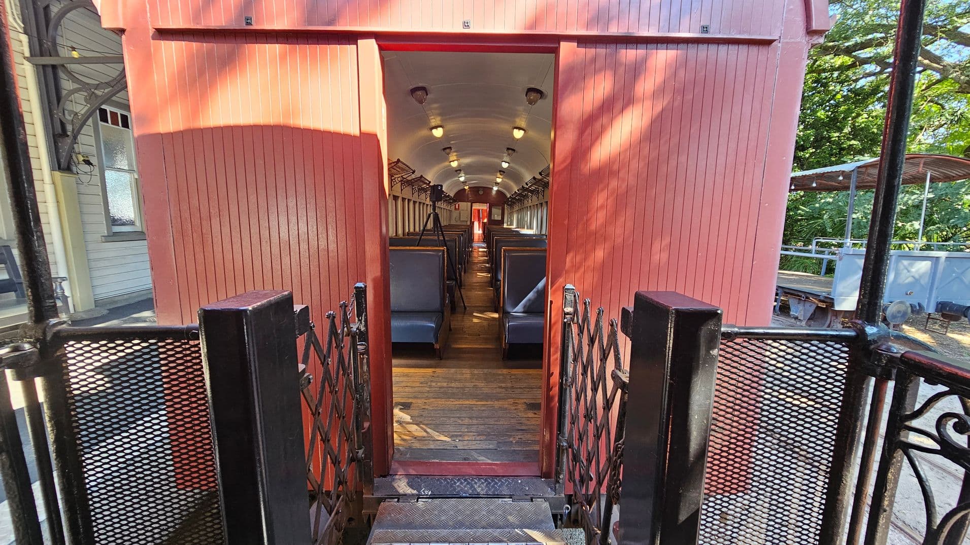 A vintage train carriage with a red exterior and wooden panelling. The entrance is open, revealing rows of blue seats inside. The ceiling is curved with small lights, and there are luggage racks above the seats. The floor is wooden. In the foreground, a black metal gate with mesh panels leads to the carriage. To the left, part of a building with a window is visible, and to the right, there are trees and a covered platform area.
