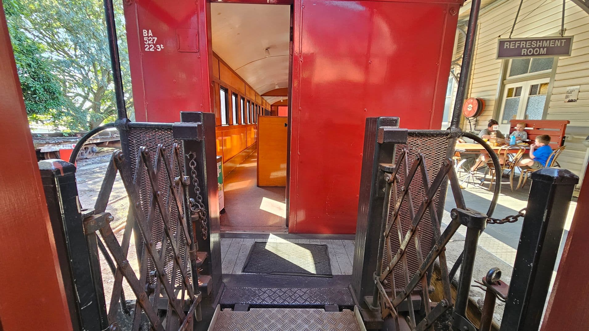 A red train carriage with an open door is in the centre, revealing a wooden interior with rows of windows. In the foreground, a metal gate with chains leads into the carriage. To the right, a building with a sign reading "Refreshment Room" is visible, where people are seated at a table. On the left, there are trees and part of another train. The floor is metal and wooden, and the ceiling inside the carriage is curved and light-coloured.