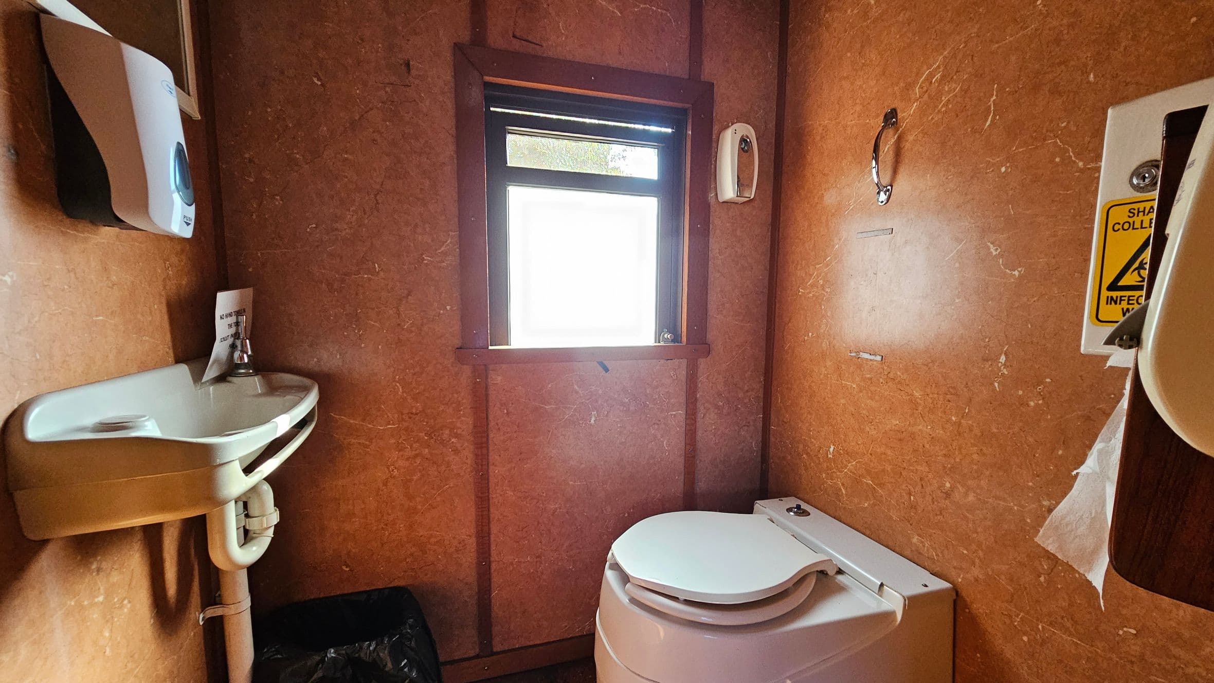 A small bathroom with brown walls. On the left, a white sink with a soap dispenser above it and a black bin below. In the centre, a window with frosted glass allows light in. On the right, a white toilet with a closed lid. Above the toilet, a metal grab handle is attached to the wall. A sharps disposal container with a warning label is partially visible on the right edge. The floor and ceiling are not visible.