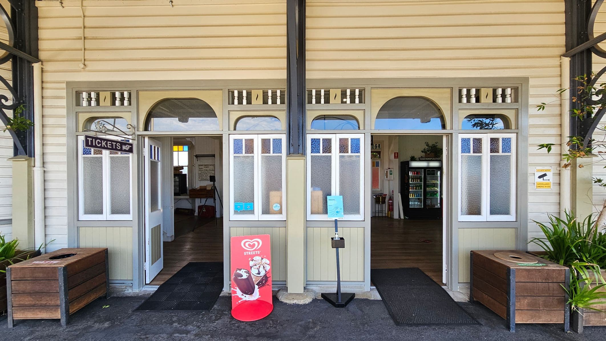 A wooden building facade with two doors and windows. The left door has a "TICKETS" sign above it. The right door is open, revealing a room with a vending machine. A red ice cream advertisement stand is in front of the right door. Two wooden planters with green plants are on either side. The floor is dark and the ceiling is not visible.