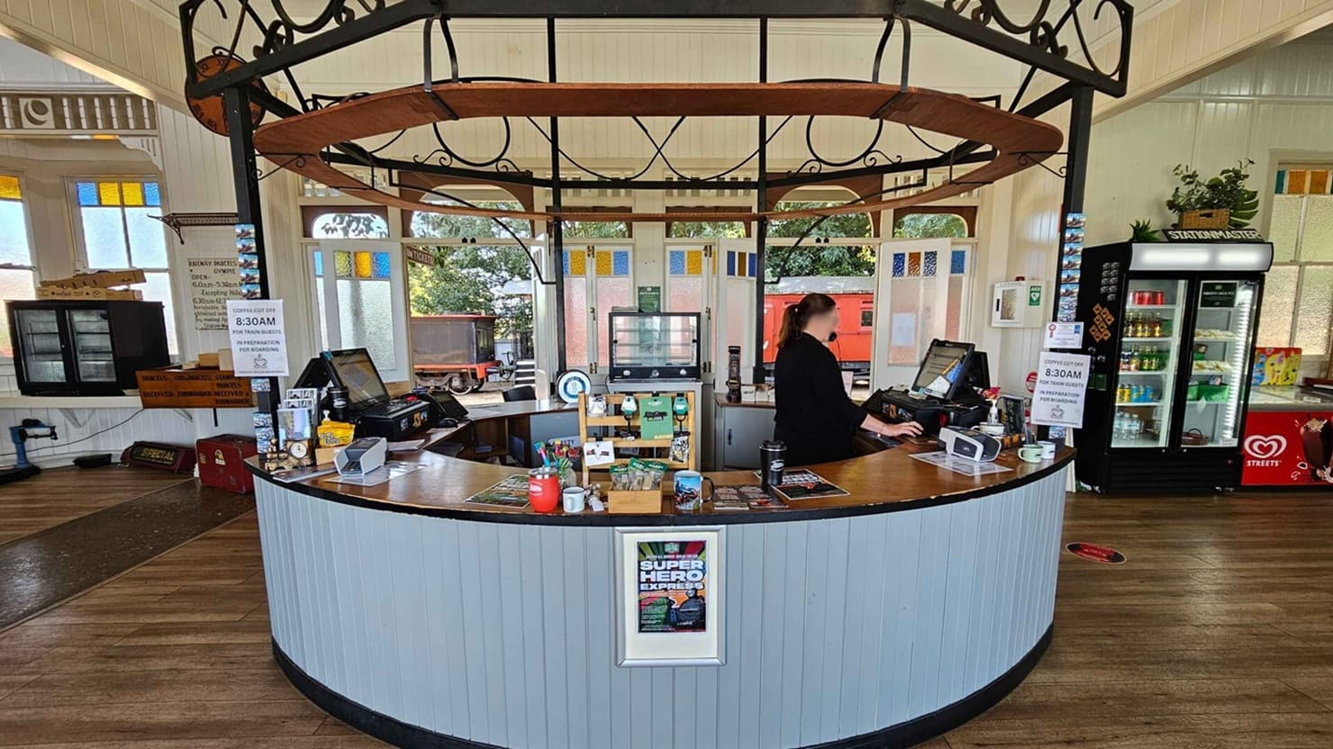A circular counter with a wooden top and light blue paneling is in the centre. A person stands behind it, working at a till. The counter displays various items, including brochures and a framed poster. To the right, a refrigerated display case holds drinks. The background features large windows with stained glass panels, and outside, a red train carriage is visible. The floor is wooden, and the ceiling has a decorative metal framework.