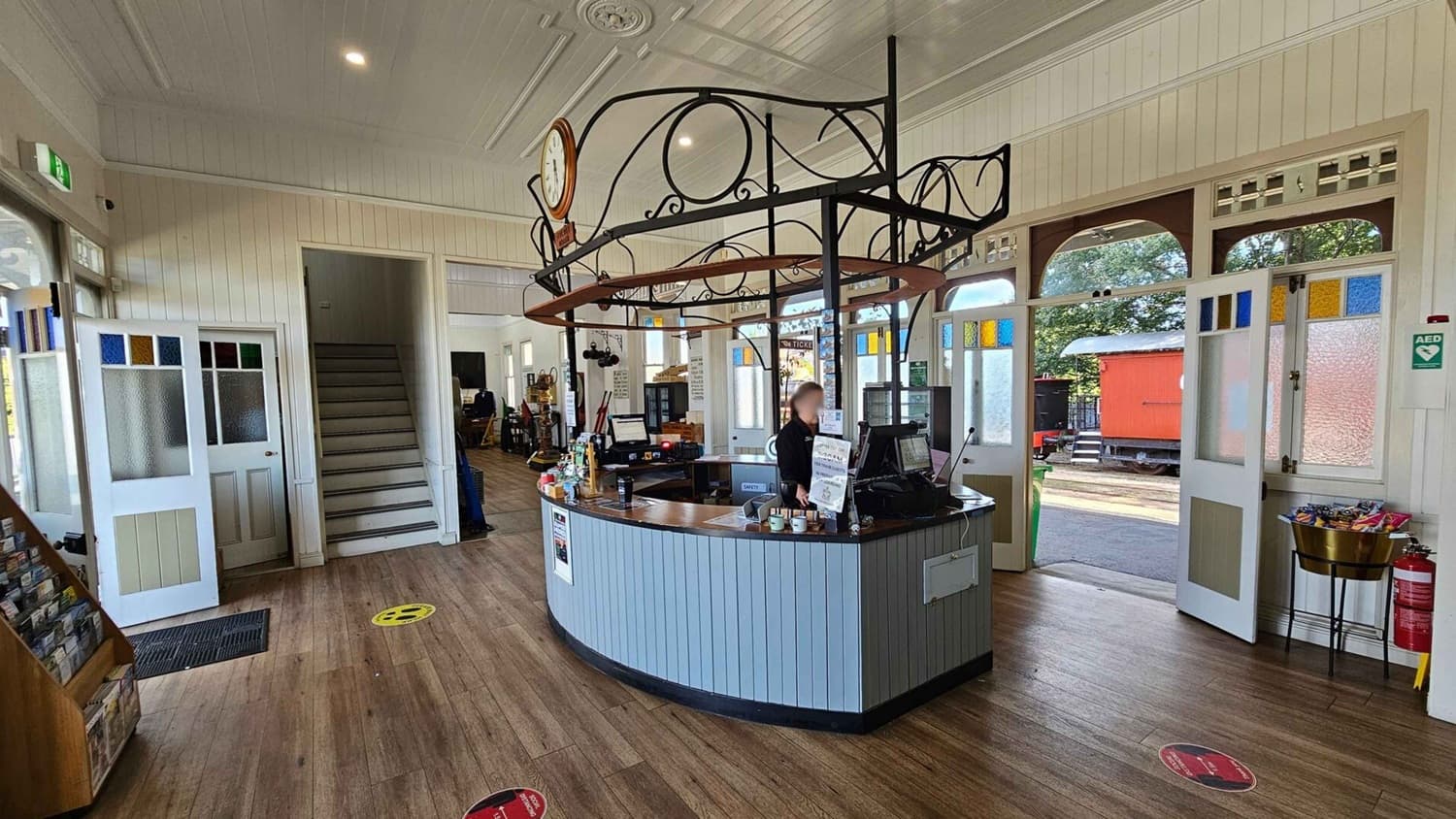 A spacious interior with wooden flooring and a light-coloured panelled ceiling. In the centre, a curved counter with a person standing behind it, working at a computer. Above the counter is a decorative metal structure with a clock. To the left, a staircase leads upwards, and a door with stained glass panels is open. On the right, double doors with colourful glass panes open to an outdoor area with a red train carriage visible. A fire extinguisher and a small snack display are nearby.