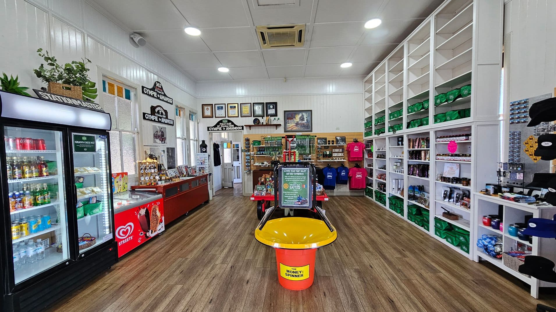 A shop interior with a wooden floor and a white panelled ceiling. On the left, a refrigerated display case holds drinks and snacks. Next to it, a red ice cream freezer is visible. The back wall features framed pictures and merchandise, including colourful shirts. On the right, tall white shelves are stocked with green bags and various products. In the centre, a yellow and red donation spinner is placed on the floor.
