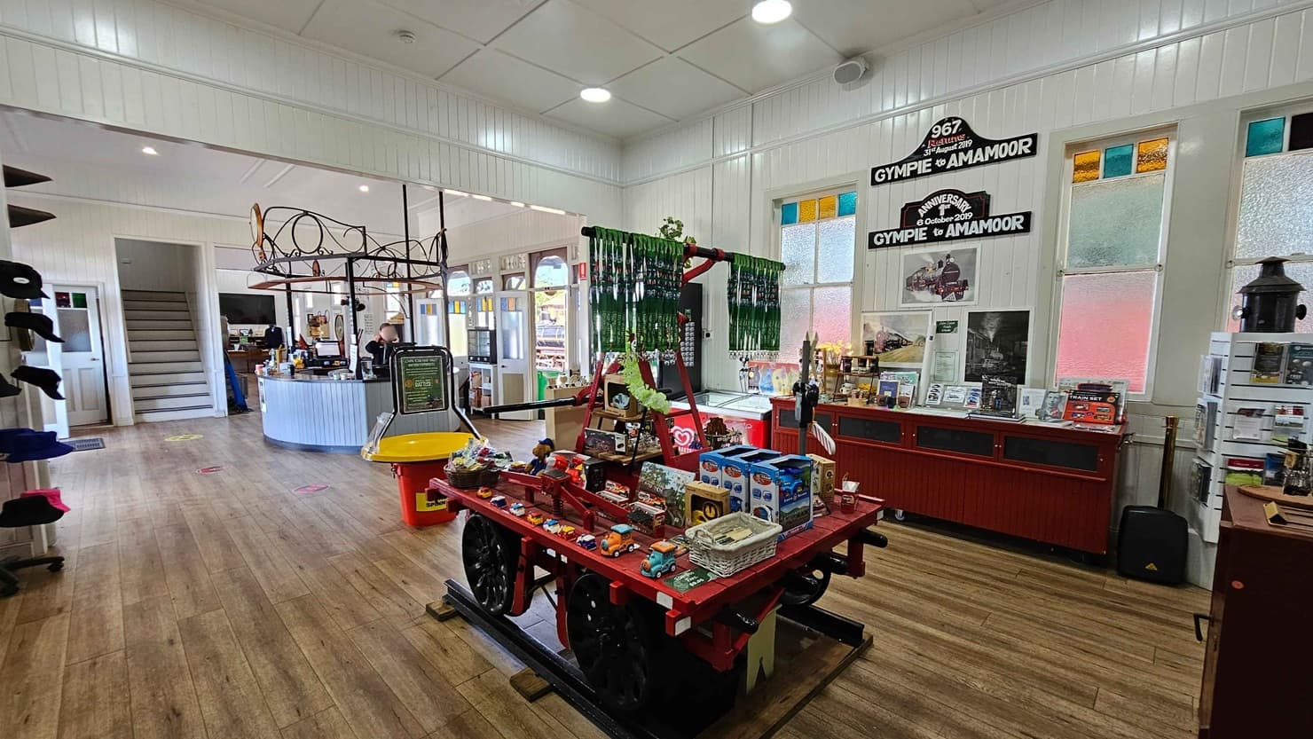 A shop interior with a wooden floor and white panelled walls. In the centre, a red cart displays various toys and souvenirs. To the right, a red counter holds more items, with signs above referencing "Gympie to Amamoor." Stained glass windows are behind the counter. On the left, a curved counter with a person behind it is near a staircase. The ceiling is white with recessed lighting.