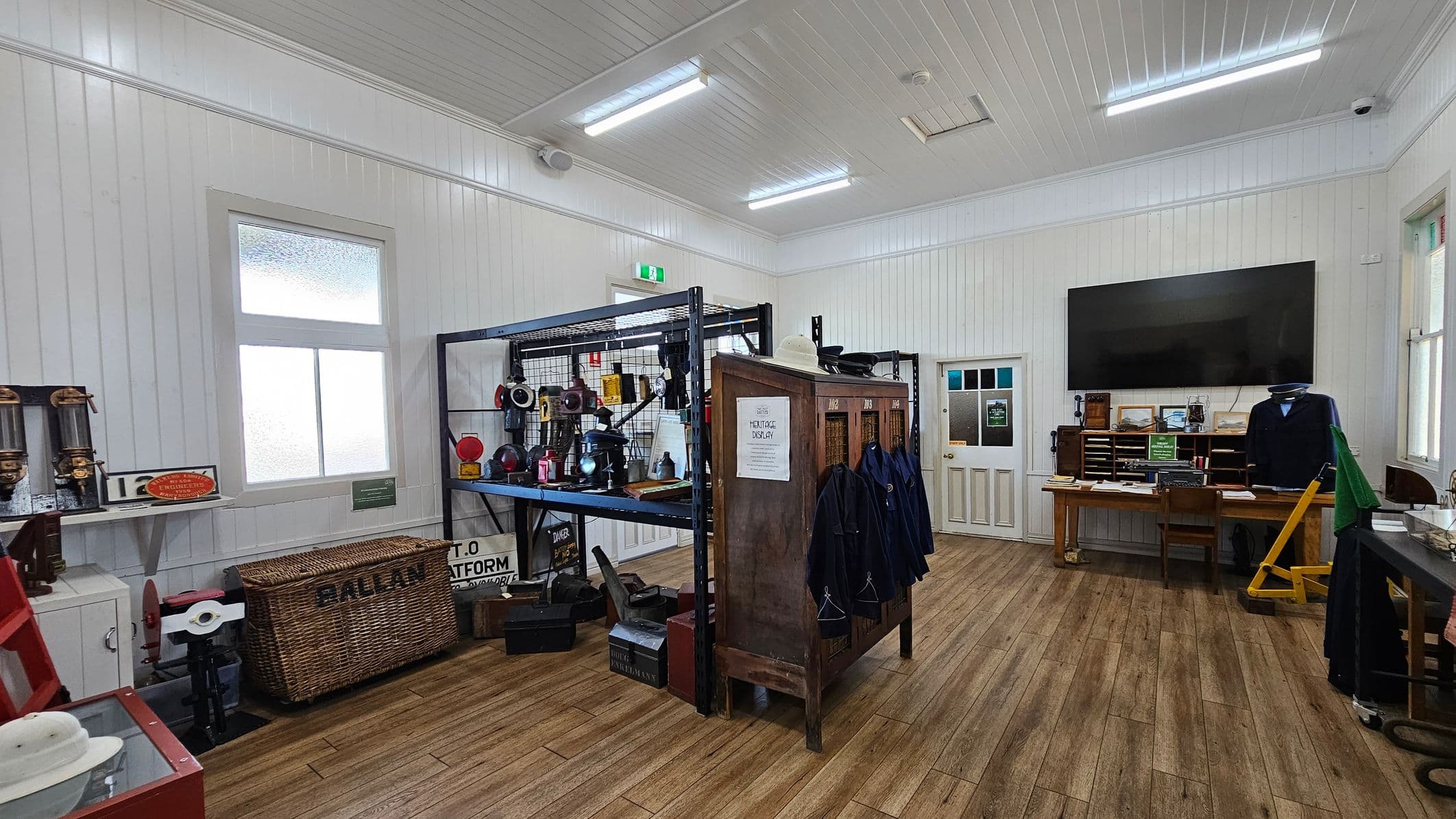 A room with white panelled walls and a wooden floor. On the left, a shelf holds vintage items, including a wicker basket labelled "BALLAN." In the centre, a metal rack displays various objects, and a wooden cabinet with coats hanging is labelled "HERITAGE DISPLAY." On the right, a large black screen is mounted above a wooden desk with office supplies. The ceiling is white with fluorescent lights, and there are windows on both sides.
