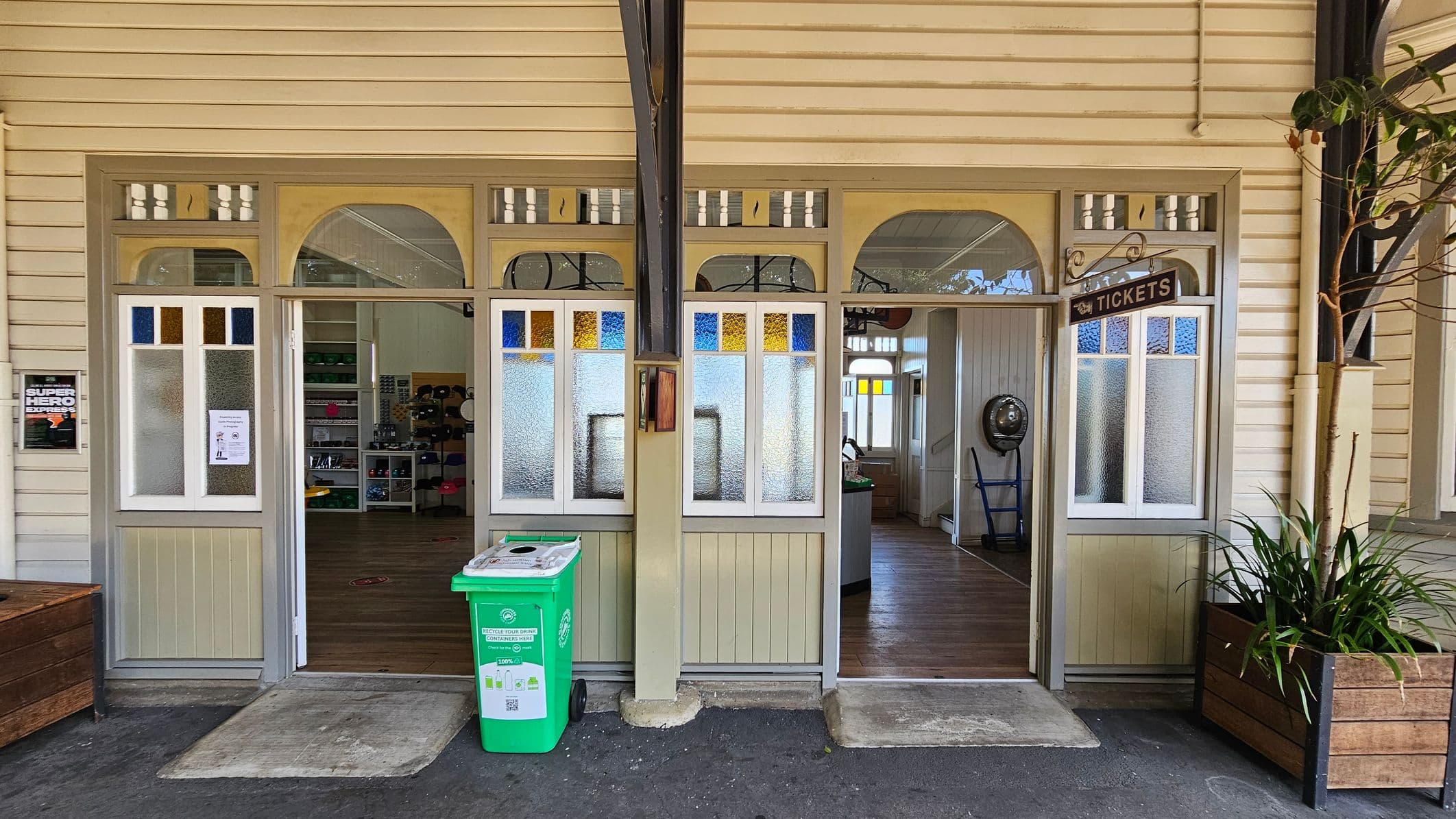 A vintage-style ticket office with a wooden exterior and frosted glass windows featuring blue and yellow stained glass accents. The entrance is open, revealing a wooden floor inside. A green recycling bin is placed outside on the left, and a "Tickets" sign hangs above the right entrance. A potted plant in a wooden planter is on the right. The ceiling is wooden, matching the exterior walls.