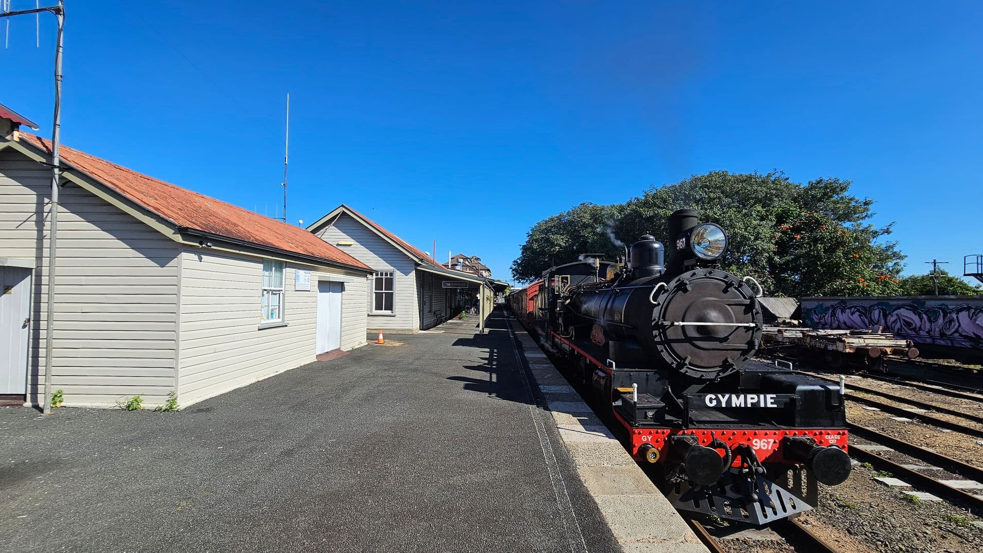 A vintage steam locomotive is on the right, labelled "GYMPIE" with the number 967. It is stationed on railway tracks beside a platform. On the left, there is a light-coloured wooden building with a red roof, featuring windows and a door. The platform is paved, and a small orange traffic cone is visible near the building. In the background, there are trees and additional railway tracks with some graffiti on a wall.