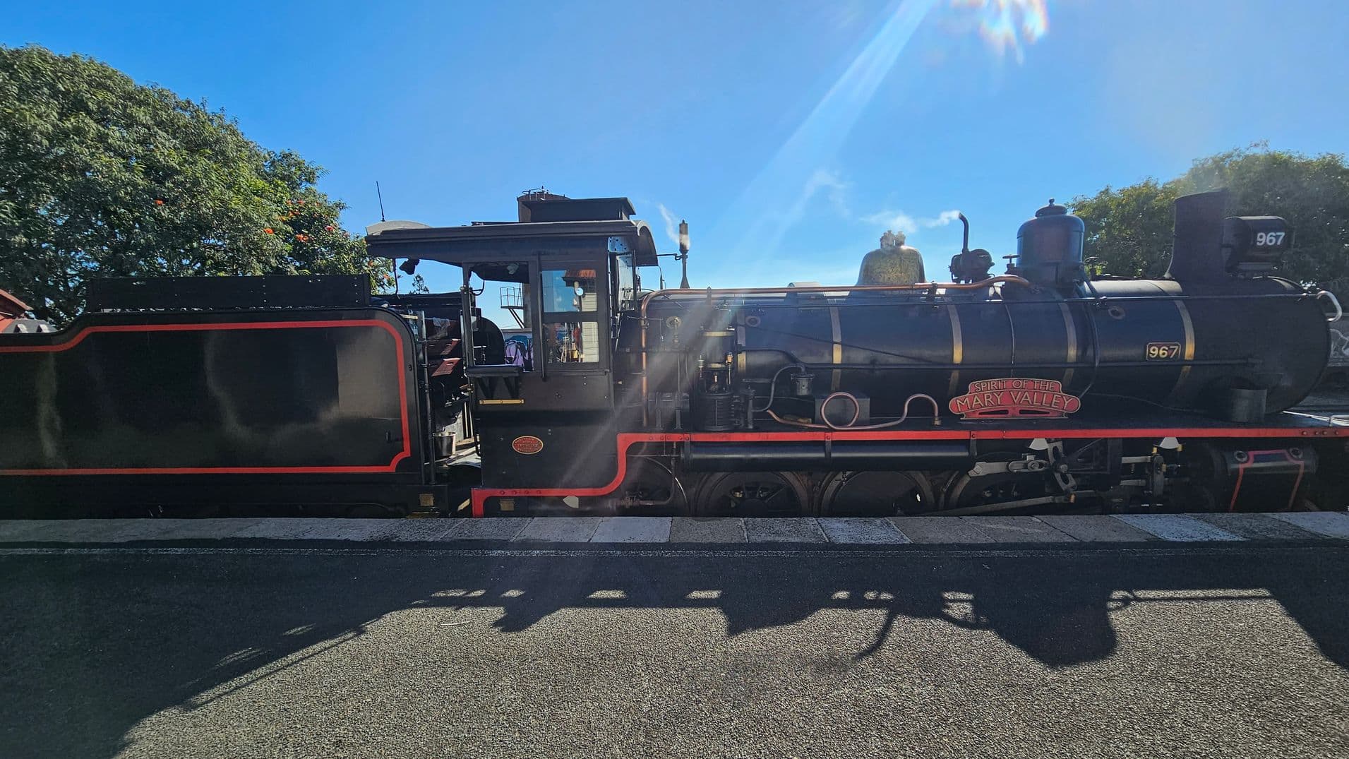 A vintage steam locomotive with the name "Spirit of the Mary Valley" is on a railway track. The train is black with red and gold detailing. The number "967" is visible on the front and side. In the background, there are trees with green foliage and some red flowers. The sky is clear and blue, and sunlight casts shadows on the ground. The platform is made of concrete with a textured surface.