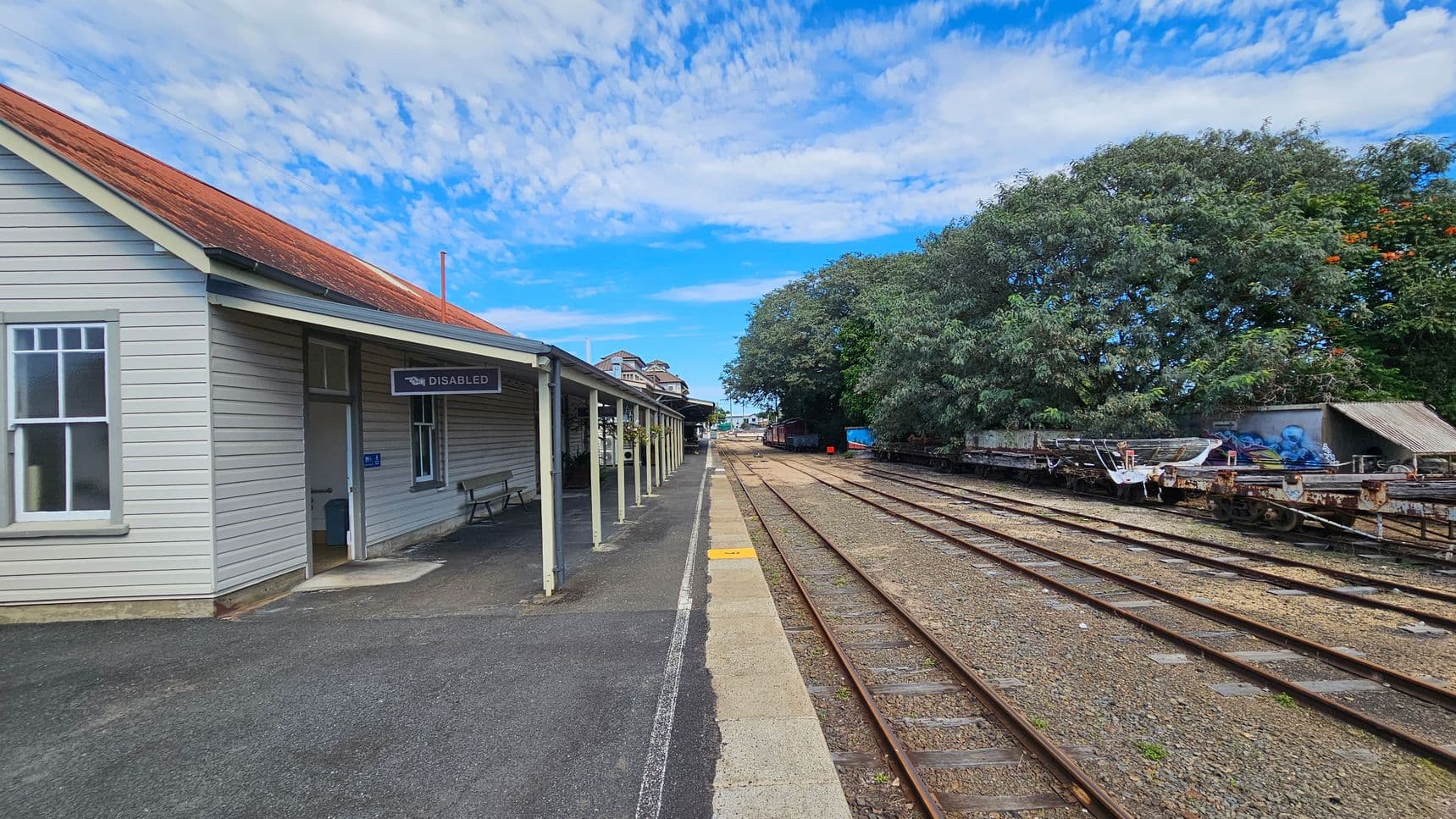 A railway station platform with a light grey wooden building on the left, featuring a red roof and a sign marked "DISABLED". A bench is positioned under the building's overhang. The platform extends into the distance, bordered by multiple railway tracks on the right. Beyond the tracks, there are trees with dense green foliage and some orange flowers. Several flatbed rail cars are visible, with some covered items. The sky is partly cloudy.
