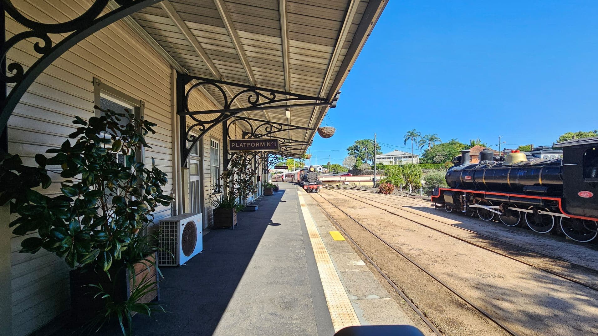 A vintage train station platform with a wooden structure on the left, featuring decorative metal brackets and a "Platform No 1" sign. Potted plants and an air conditioning unit are placed along the wall. On the right, a black steam locomotive is on the tracks. The background includes trees, a house, and a clear blue sky. The platform has a grey surface with a yellow tactile strip near the edge.
