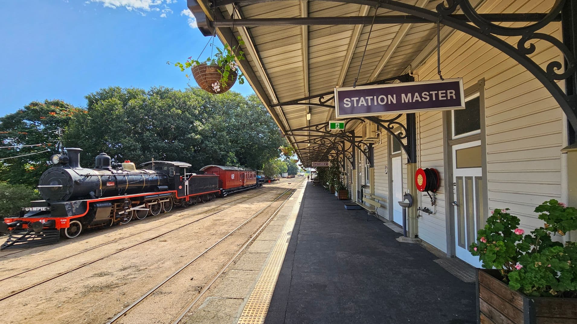 A vintage steam locomotive is on the left, with a black engine and red accents, positioned on railway tracks. On the right, a train station platform features a "Station Master" sign, a fire hose reel, and potted plants. The platform is covered by a metal roof with decorative brackets. A hanging basket with greenery is suspended from the roof. The background includes lush green trees and a clear view down the tracks.