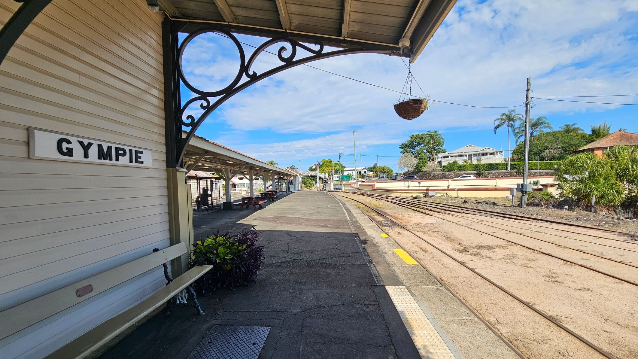 A railway station platform with a sign reading "GYMPIE" on the left. A white bench is beneath the sign, with some plants nearby. The platform extends into the distance, with a covered waiting area and seating. Train tracks run parallel to the platform on the right. In the background, there are houses, trees, and a clear sky. A decorative metal bracket and hanging basket are visible under the roof.