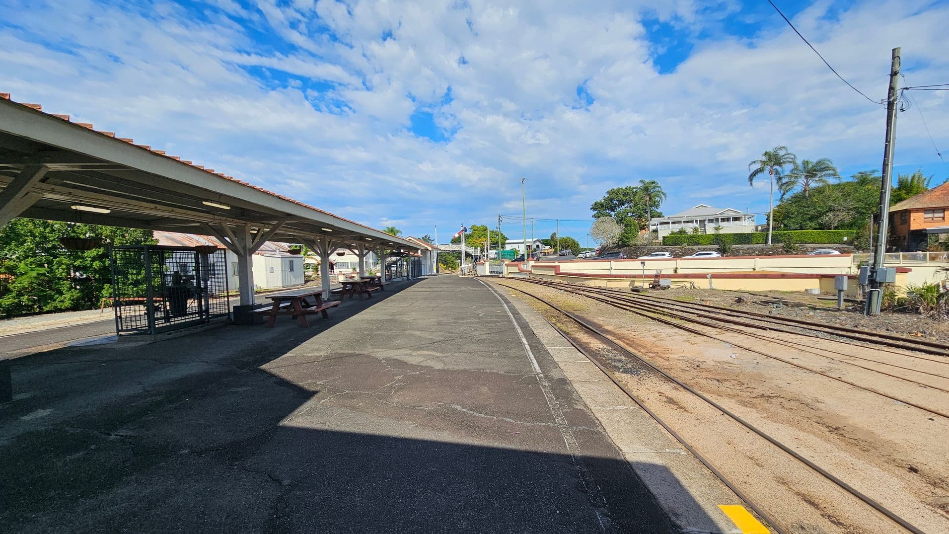 A railway station platform with a covered waiting area on the left, featuring benches and a fenced section. The platform is paved with a mix of green and brown surfaces. On the right, multiple railway tracks run parallel to the platform. In the background, there are residential buildings, trees, and a few parked cars. The sky is partly cloudy, and the area is well-lit by daylight.