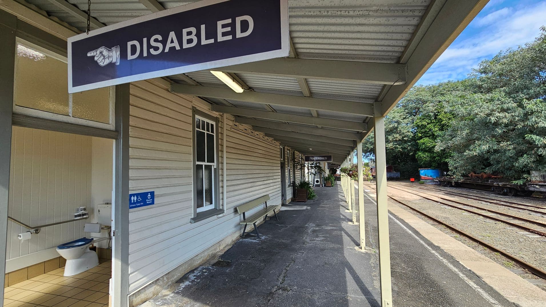 A railway station platform with a covered walkway. On the left, there's a sign reading "DISABLED" with an arrow pointing towards an accessible toilet, visible with a grab rail and toilet paper holder. The building has light-coloured wooden siding and a bench. The platform extends into the distance with a "LADIES" sign further along. On the right, railway tracks run parallel to the platform, bordered by dense green trees. The ceiling is metal, and the floor is paved.