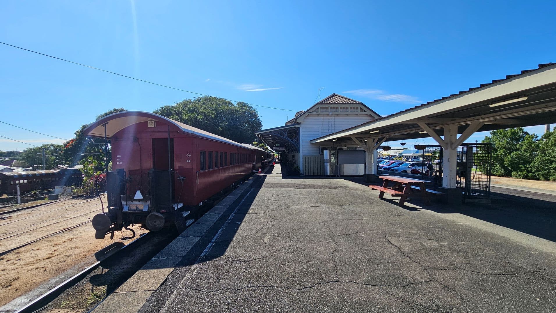 A vintage red train carriage is on the left, positioned on railway tracks. To the right, there's a covered platform with a wooden bench and a picnic table. The station building is in the background, featuring a gabled roof. The ground is paved with cracked asphalt, and there are trees and parked cars visible in the distance.
