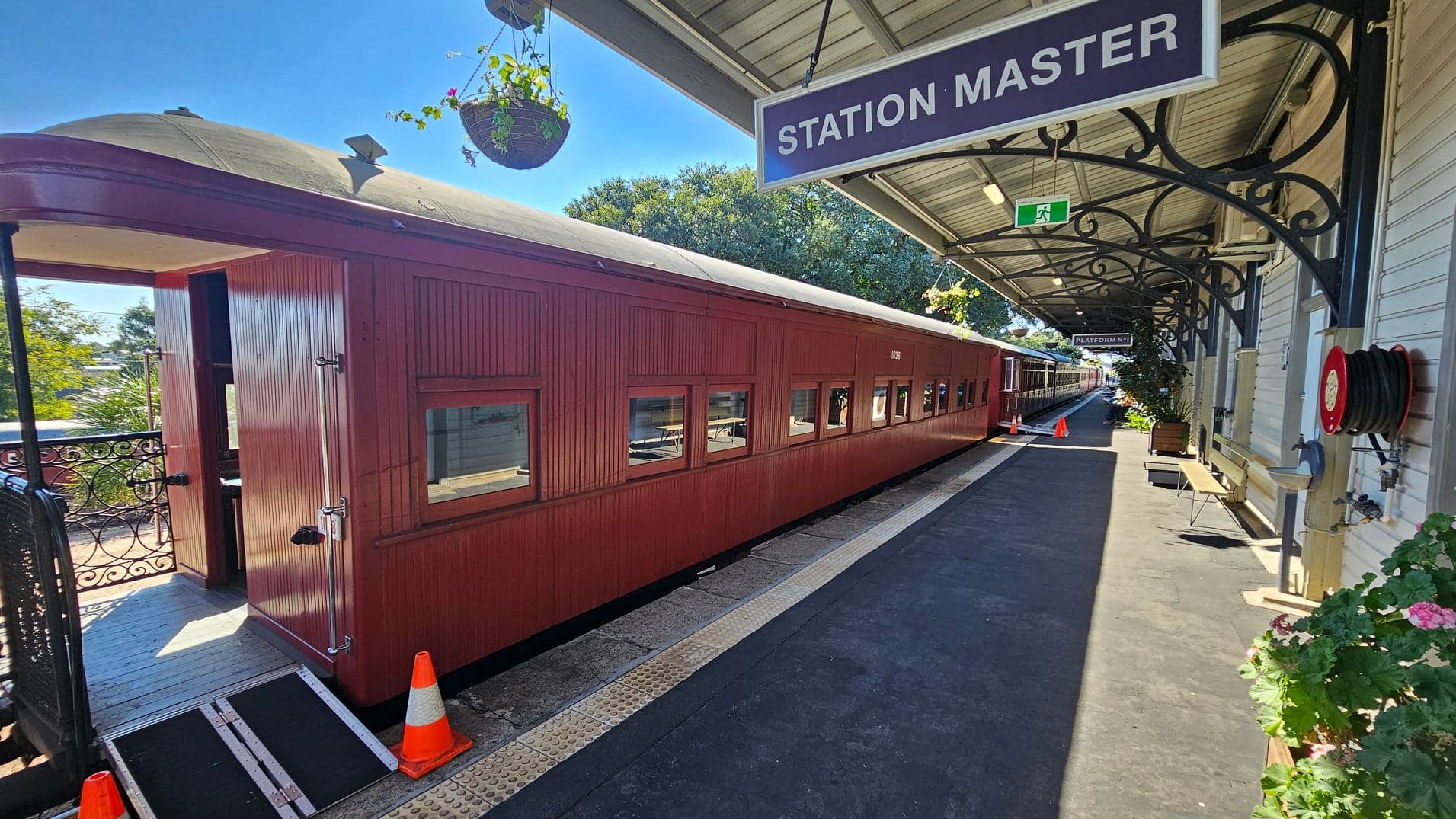 A vintage red train carriage is on the left, with a ramp and orange cones near the entrance. The platform on the right has a "STATION MASTER" sign hanging from a metal frame. The platform is lined with a green floor, a fire hose reel, and potted plants. The ceiling is covered with a metal roof, and hanging baskets with plants are suspended above. Trees are visible in the background, and a "PLATFORM 1" sign is further down the platform.