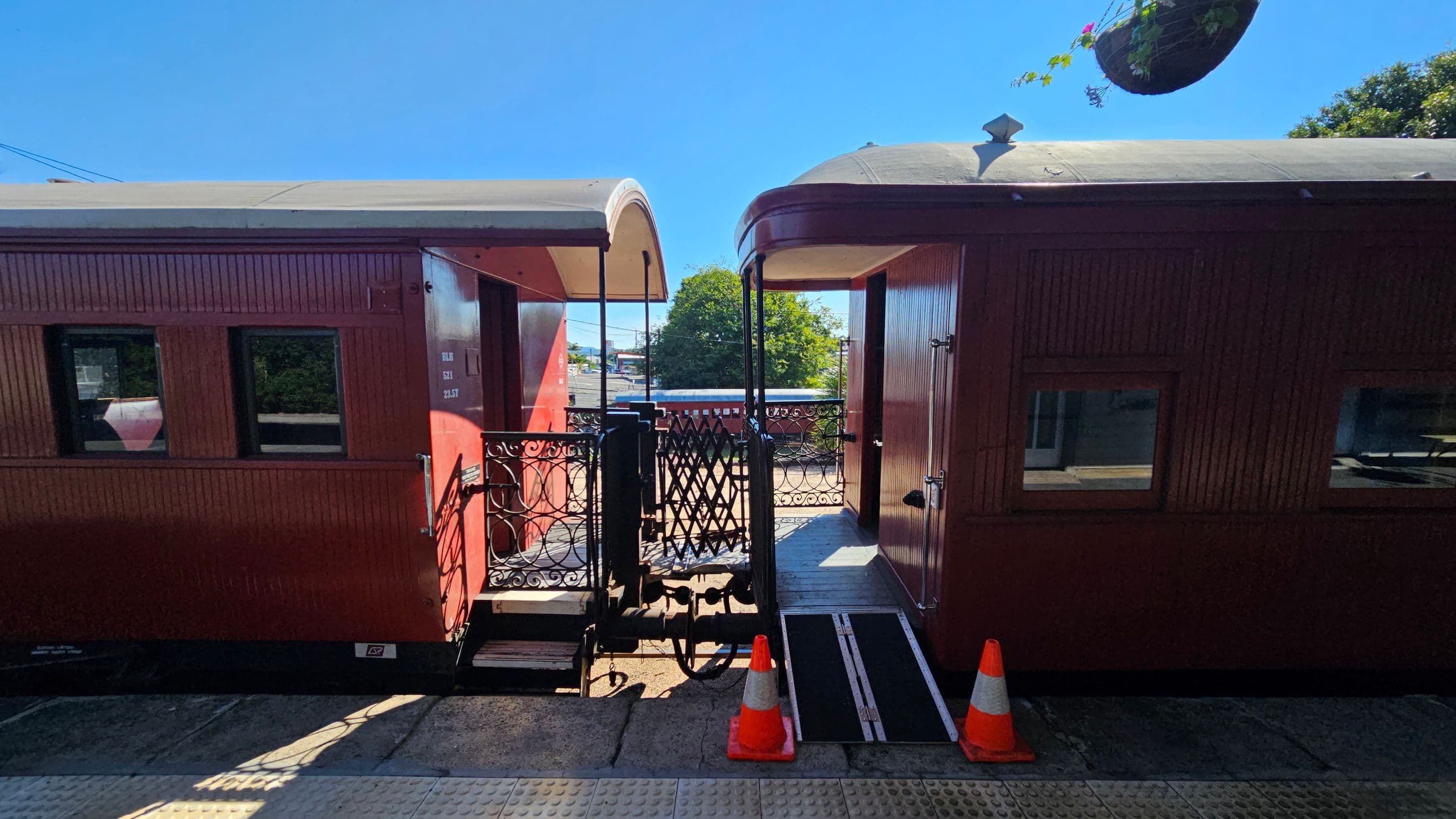 Two vintage train carriages are connected by a metal gate in the centre. The carriages are painted in a reddish-brown colour with visible windows. A ramp and two orange traffic cones are placed on the platform in front of the connection. In the background, there is greenery and a hanging plant basket. The platform surface is textured with tactile paving.