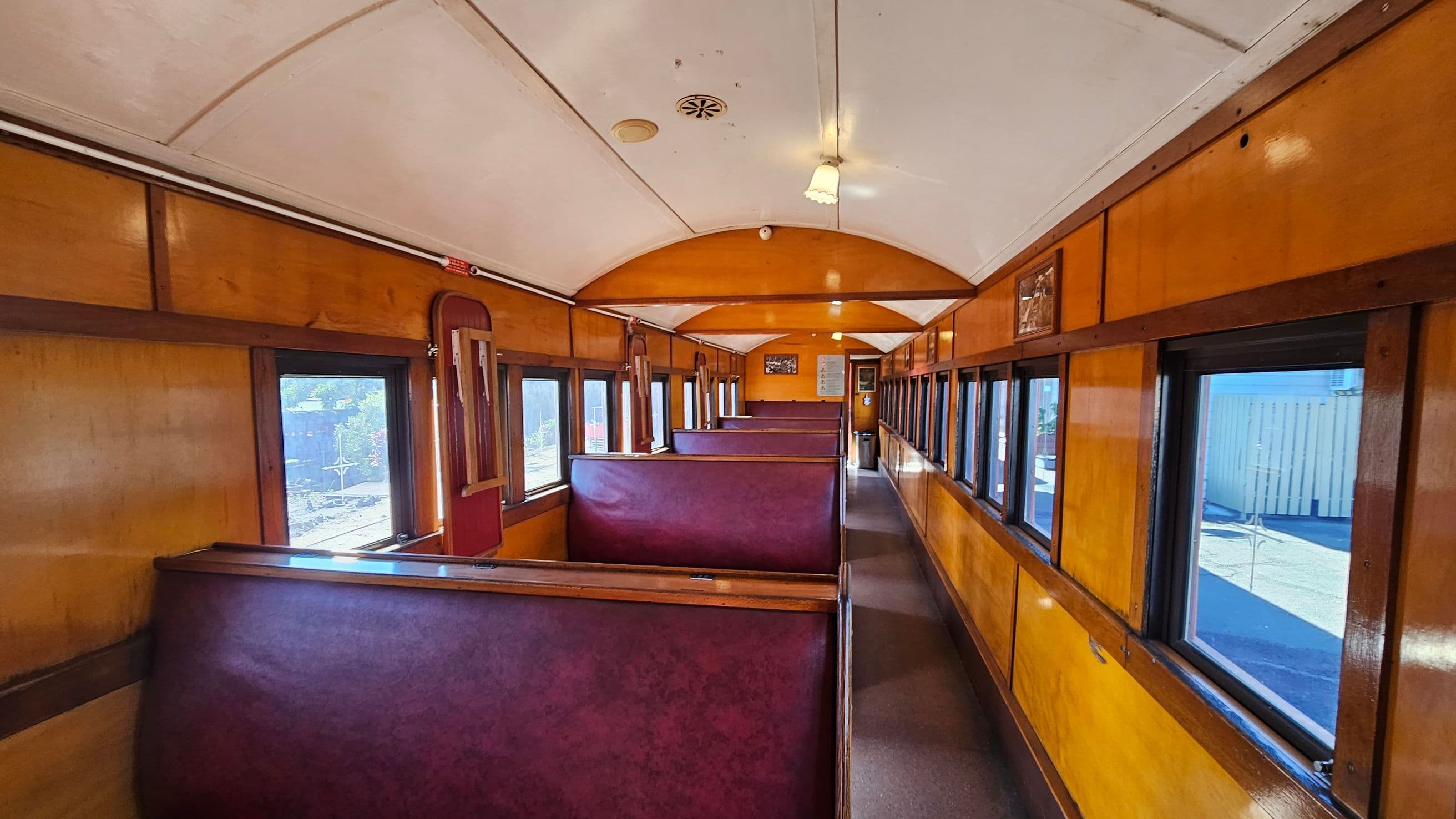 Interior of a vintage train carriage with wooden panelling and maroon upholstered bench seats. The ceiling is curved and painted white, with small light fixtures. Large rectangular windows line both sides, allowing natural light to illuminate the space. The floor is carpeted in a dark colour. At the far end, there are signs and a door. The overall atmosphere is classic and nostalgic.