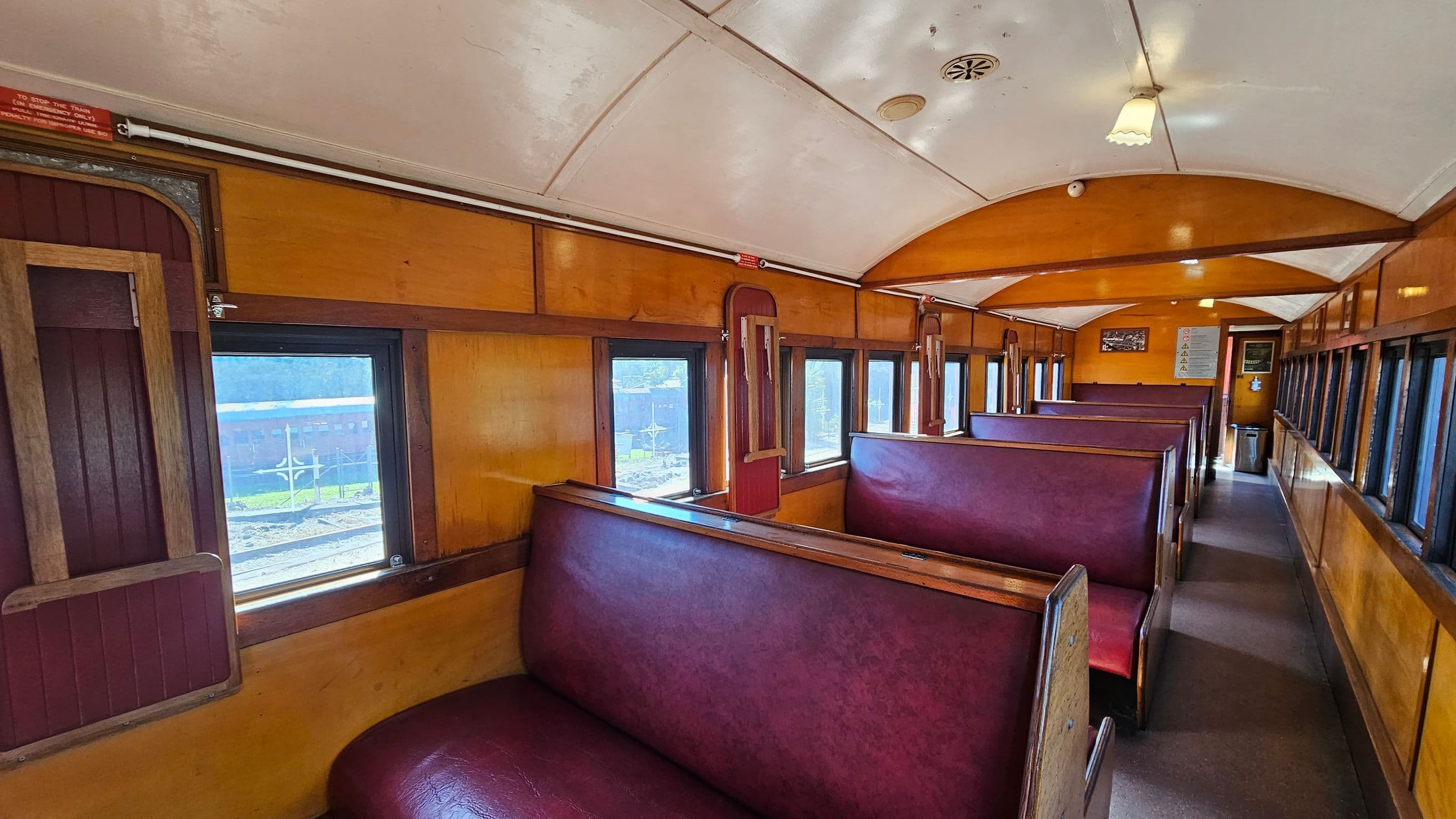 Interior of a vintage train carriage with wooden panelling and maroon leather bench seats. On the left, a series of windows with wooden frames allow natural light to enter. The ceiling is arched and painted white, with small light fixtures. The floor is carpeted in a neutral tone. Towards the background, a door and some signage are visible, along with more seating. The overall atmosphere is classic and nostalgic.