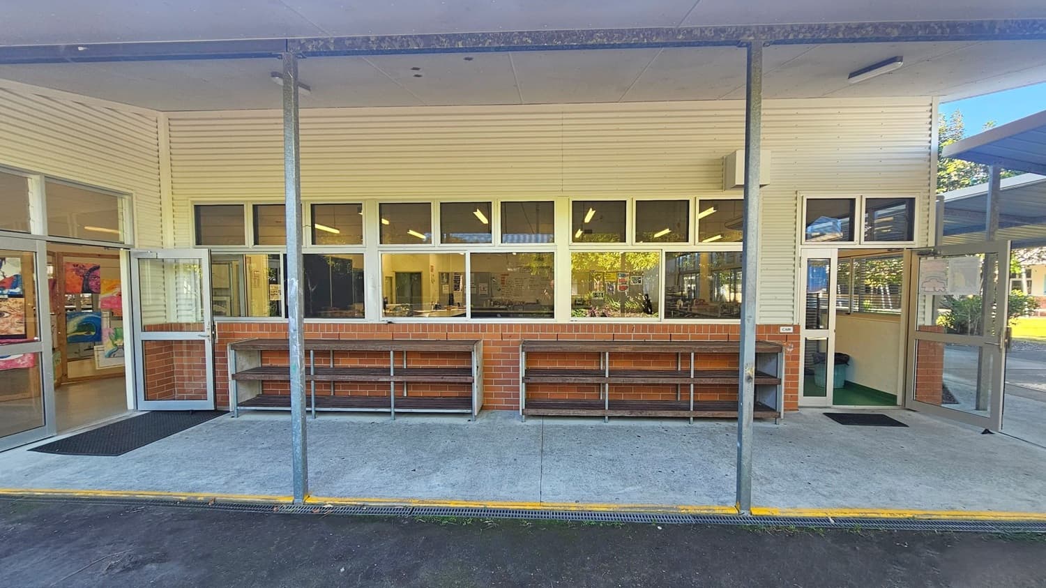 A building exterior with a brick and cream-coloured facade. In the centre, there are large windows with a view into a room with tables and chairs. Below the windows, there are metal and wood shelves. On the left and right, there are open glass doors leading inside. The floor is concrete with a black mat near the doors. The ceiling is plain and white, supported by metal poles. Outside, there is a dark asphalt surface with some leaves scattered.
